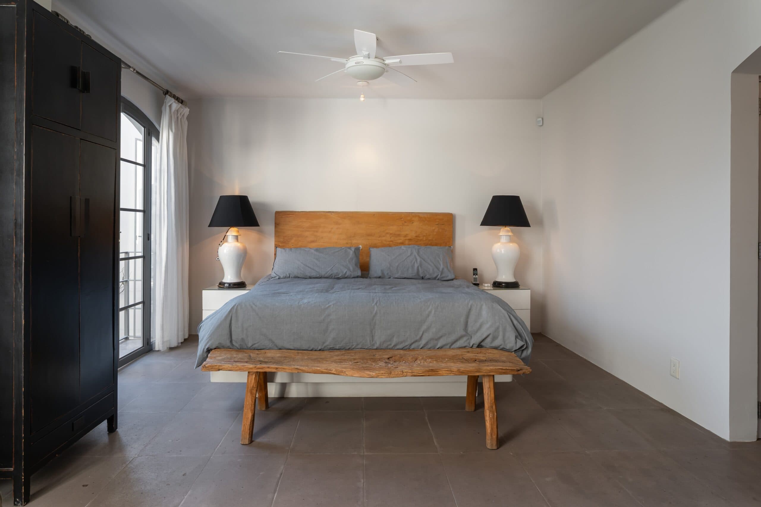Bedroom with gray bedding, wooden headboard, matching nightstands and lamps on each side of the bed; a rustic wooden bench at the foot and a dark wardrobe on the left, San Miguel de Allende