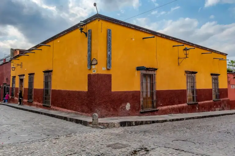 Bright yellow building with a red base and iron-barred windows along a cobblestone street, with vintage street lamps and a cloudy sky above, San Miguel de Allende