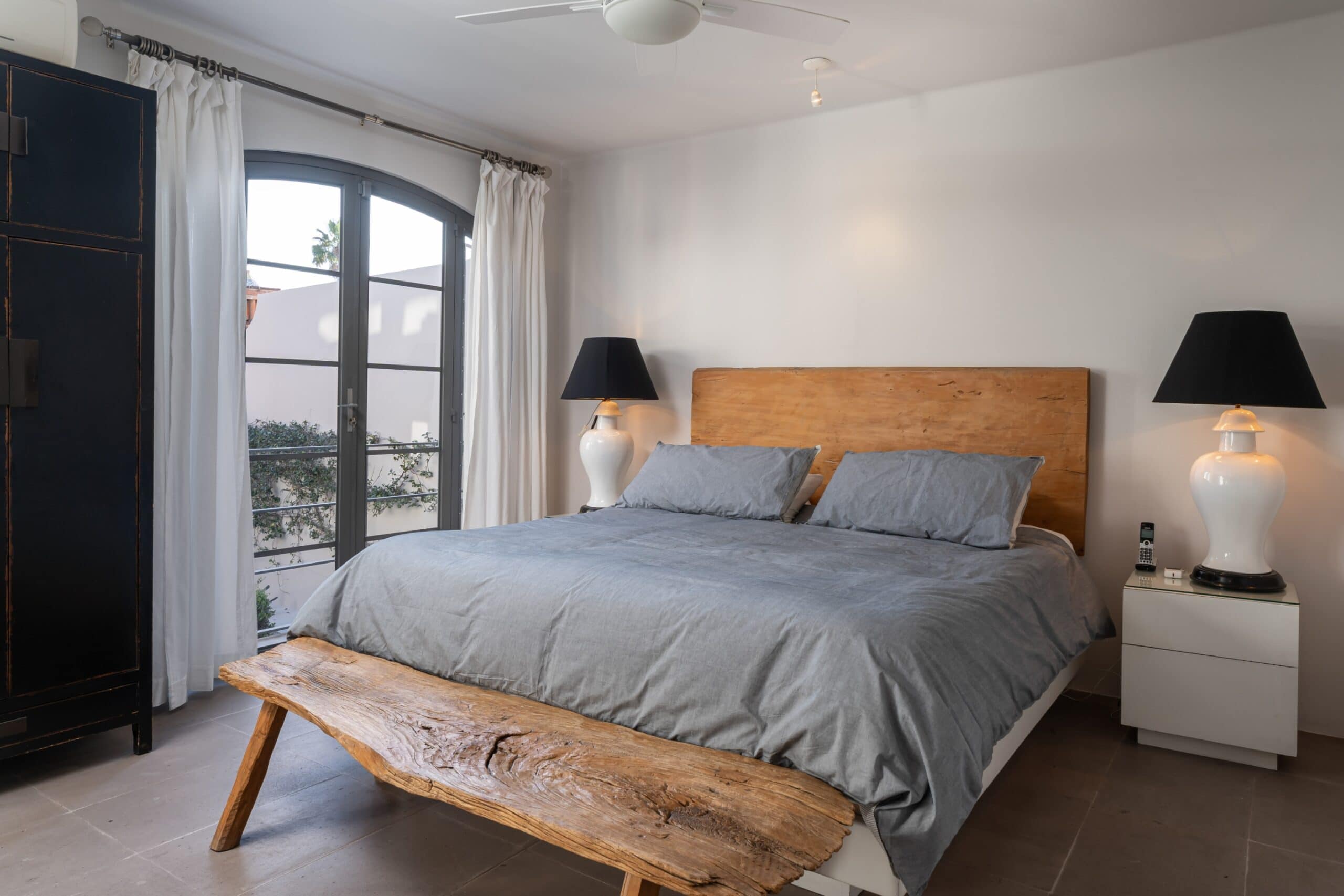 Bedroom with gray bedding, a rustic wooden bench at the foot, and a curved glass door with white curtains, San Miguel de Allende
