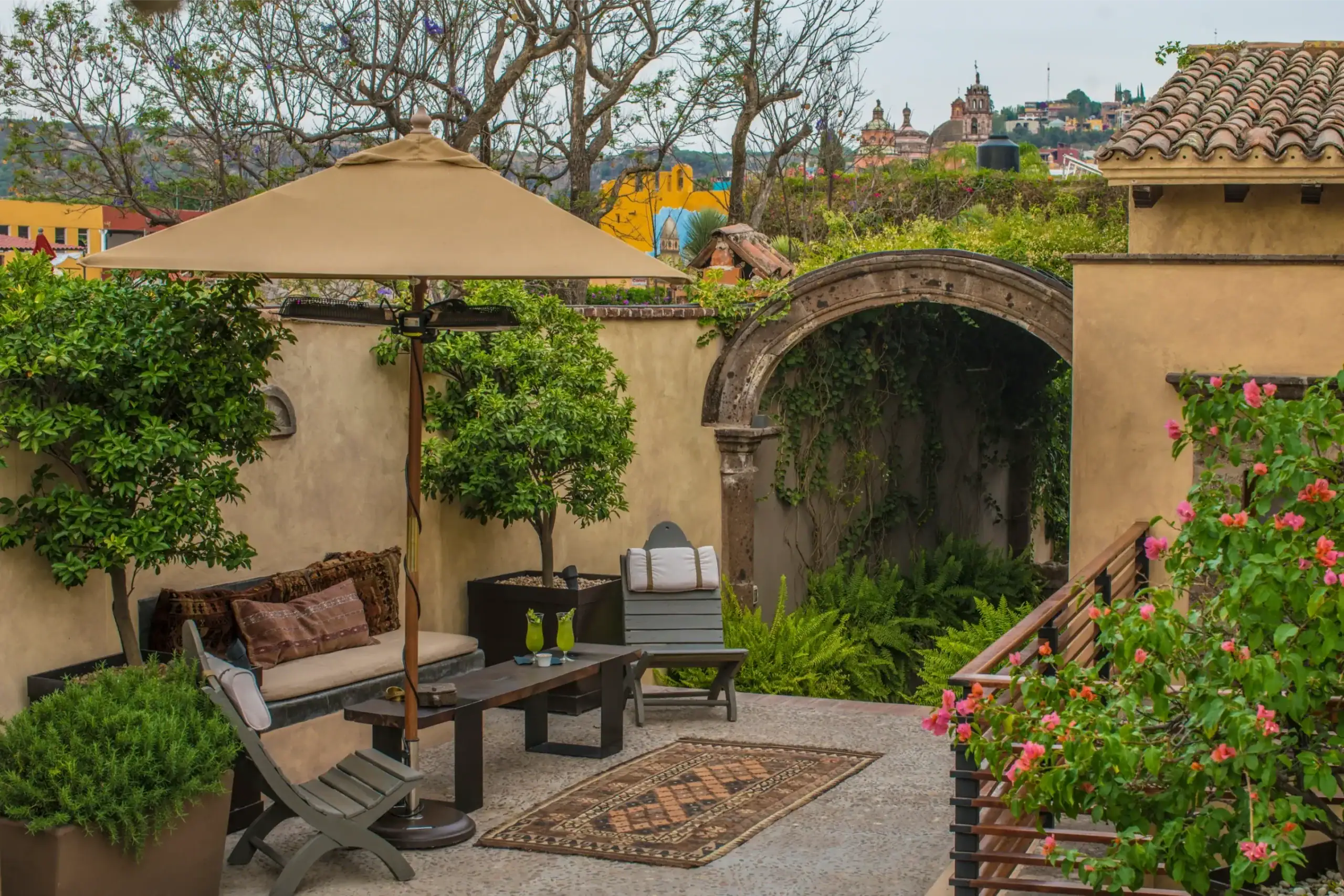 Cozy outdoor patio with a large beige umbrella, cushioned seating, and lush potted plants overlooking a colorful hillside town, San Miguel de Allende