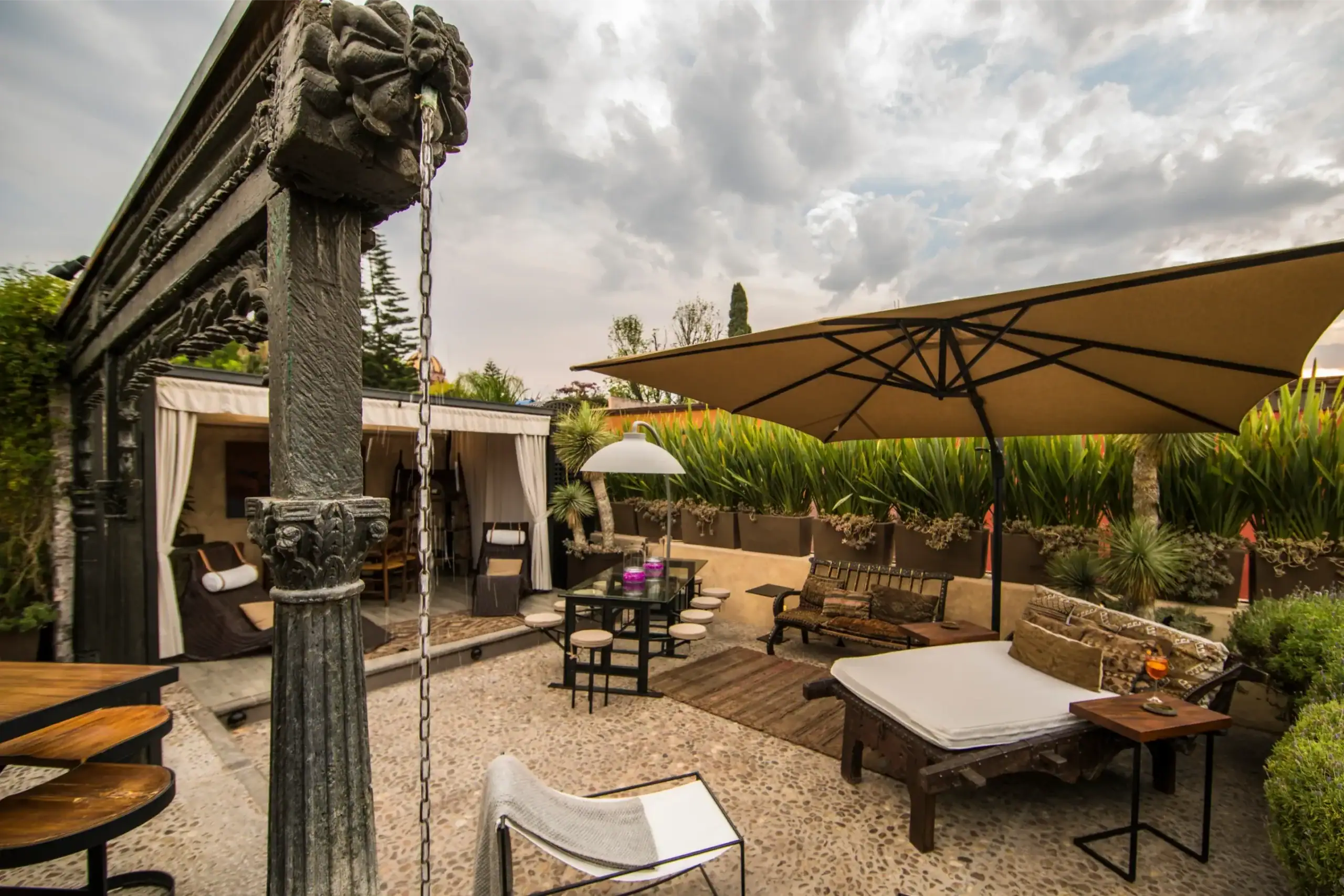 Outdoor patio with beige umbrellas, modern seating, and lush potted plants under a cloudy sky, San Miguel de Allende