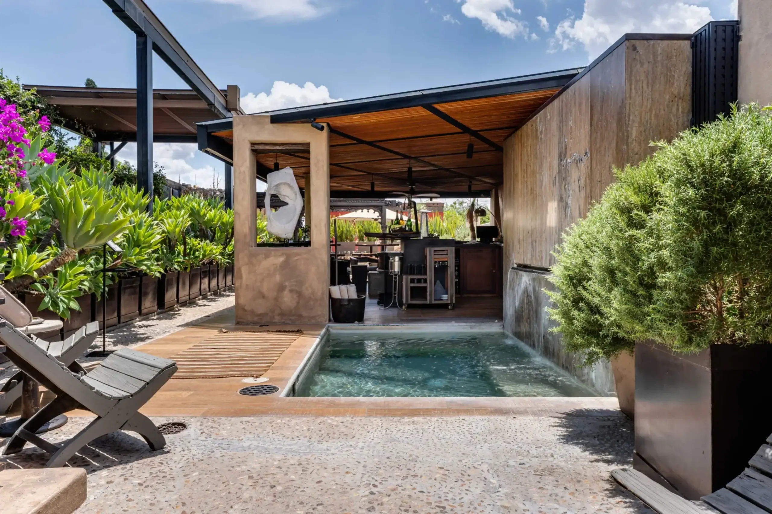 Outdoor poolside area with wooden deck, lounge chairs, and lush green plants along a border wall under a modern covered patio, San Miguel de Allende