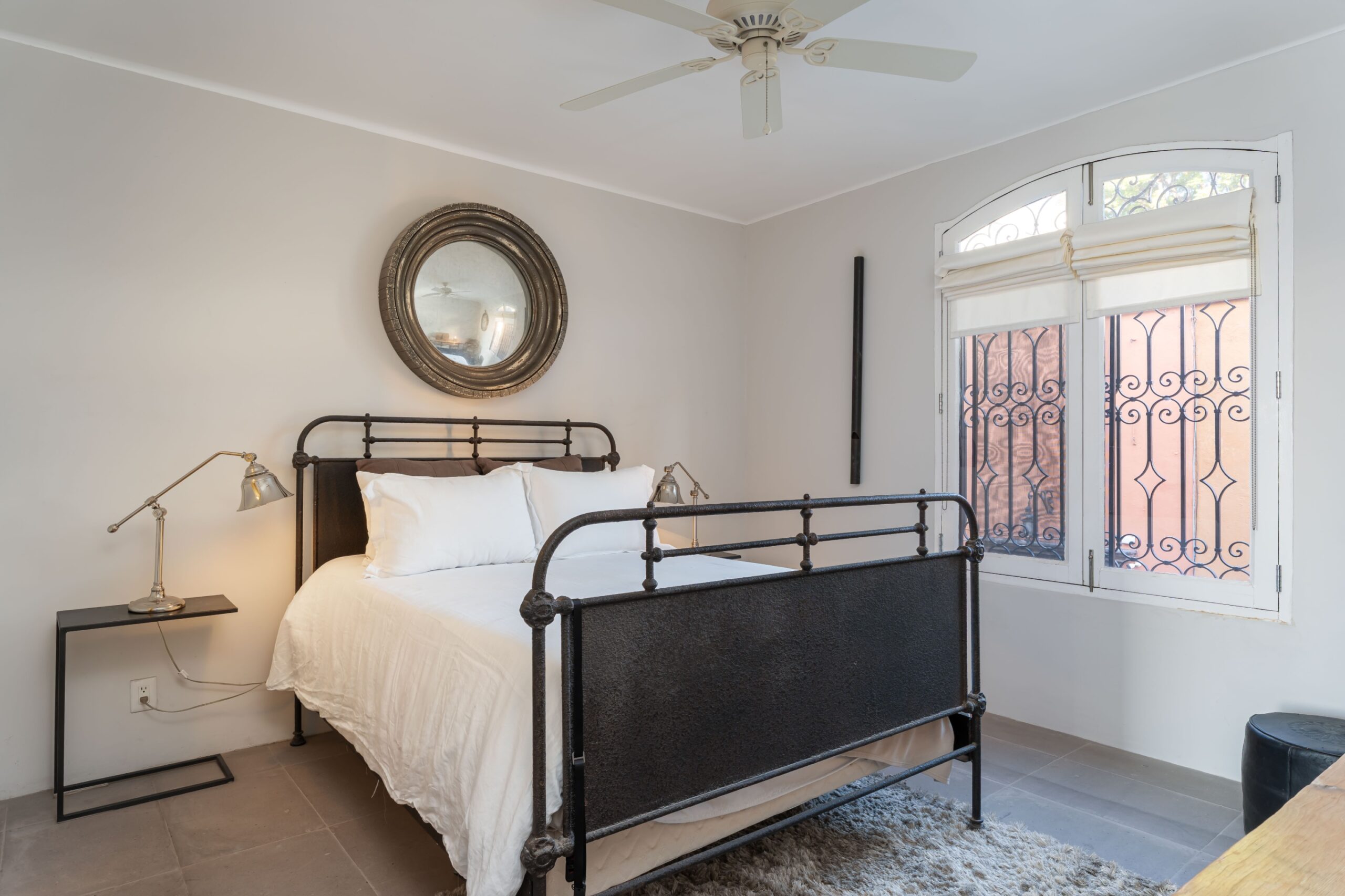 Bedroom with a black metal bed frame, white bedding, and a round decorative mirror above the headboard; a small side table lamp on the left and a large window with blinds on the right, San Miguel de Allende