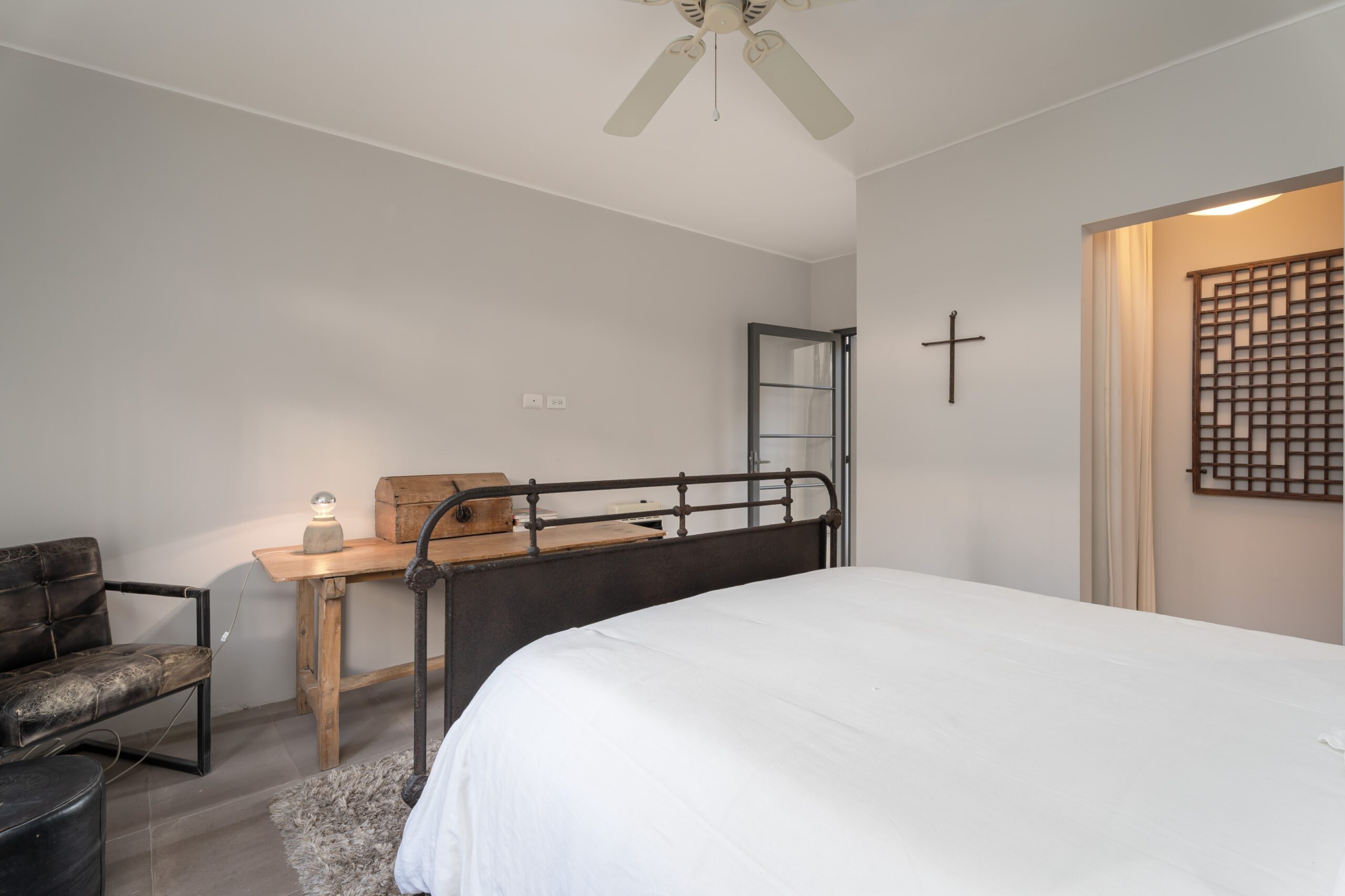 A modern bedroom with a metal-framed bed, white linens, a wooden desk with a vintage chest, and a ceiling fan above the light gray walls, San Miguel de Allende