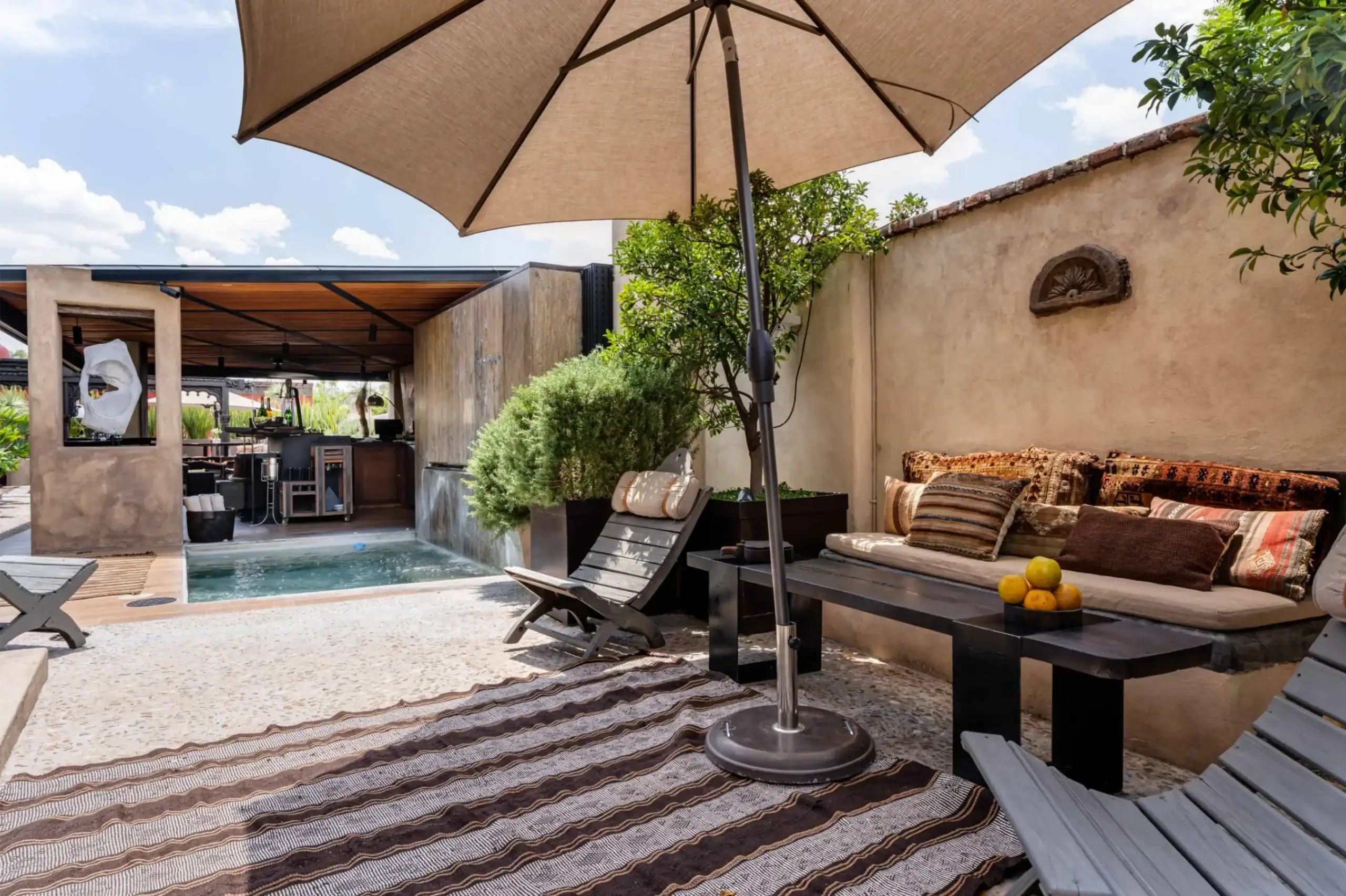 Outdoor lounge area with a large beige umbrella, low black benches, striped cushions, and a small pool bordered by a stucco wall and greenery, San Miguel de Allende