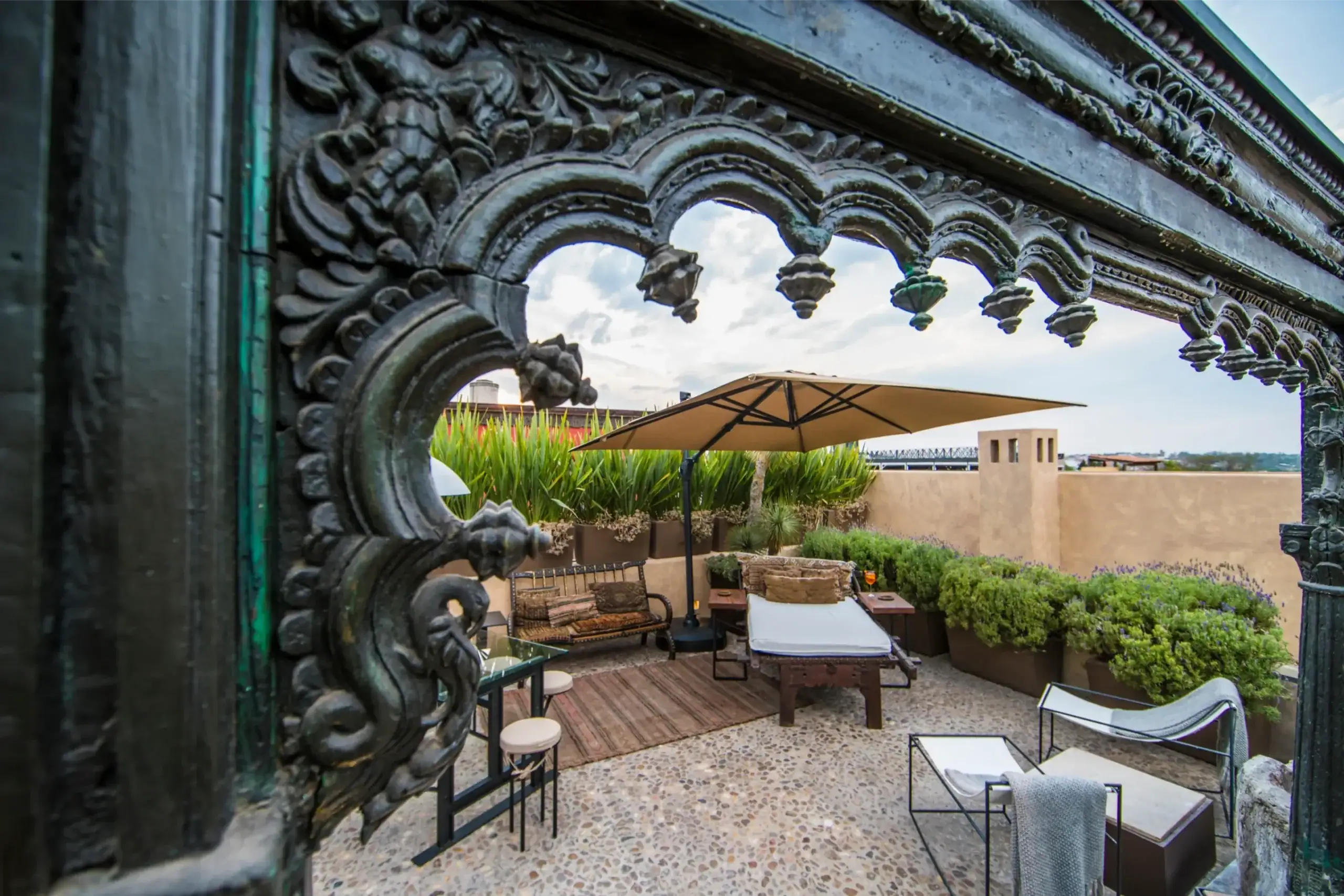 Ornate stone archway with leafy carvings opening to a sunny courtyard with a beige wall, umbrella, and outdoor seating ahead, San Miguel de Allende