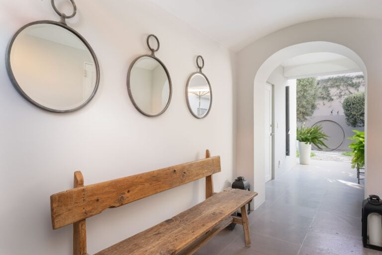 Wall hallway with three circular metal-framed mirrors above a rustic wooden bench; arched doorway opens to a courtyard with plants outside, San Miguel de Allende