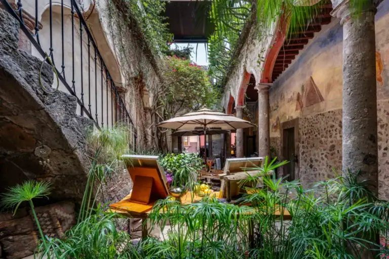 Stone-walled courtyard with arches, lush plants, and a wooden dining table shaded by a beige umbrella under a pergola collectives, San Miguel de Allende