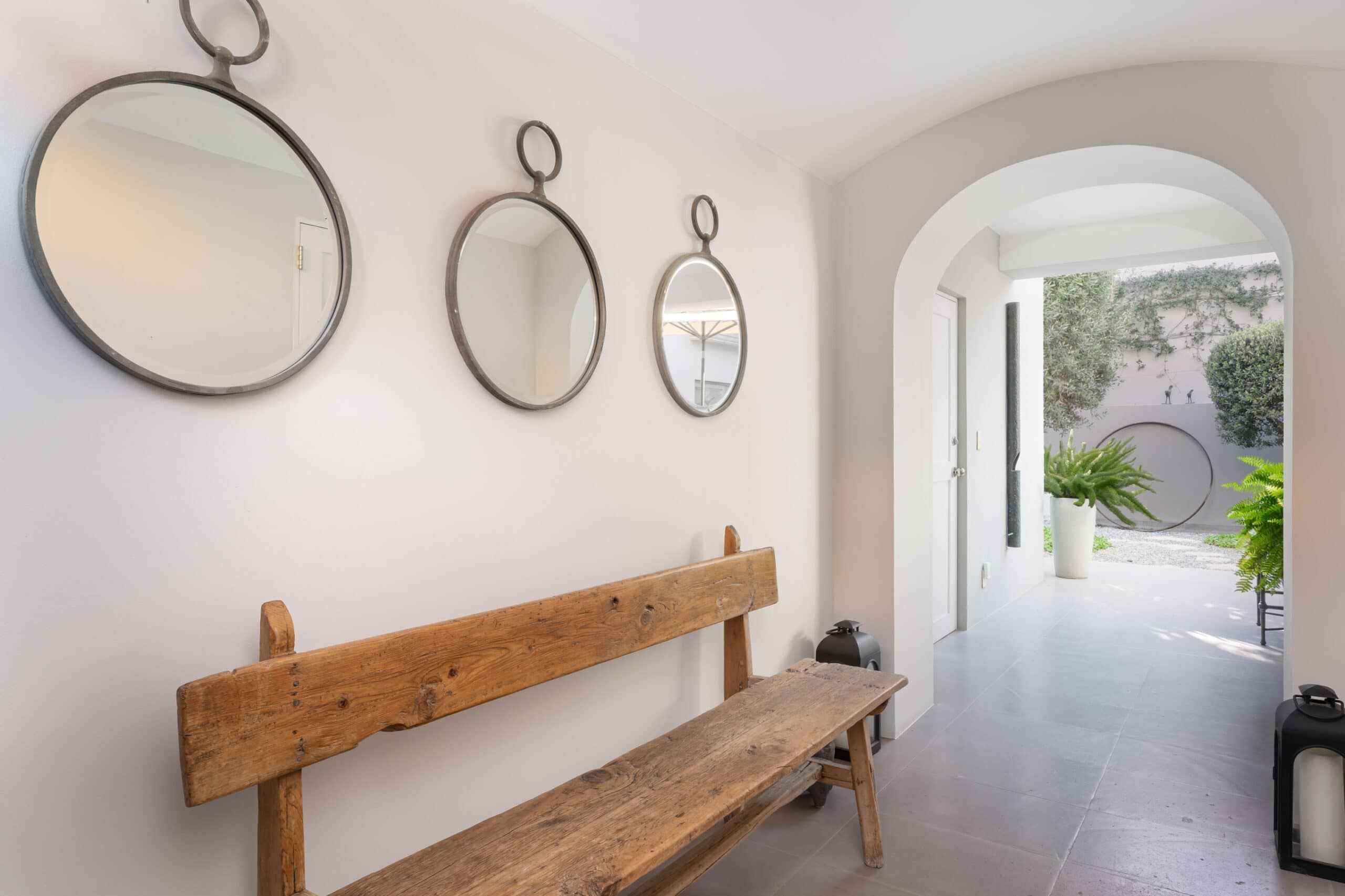 Wall hallway with three circular metal-framed mirrors above a rustic wooden bench; arched doorway opens to a courtyard with plants outside, San Miguel de Allende
