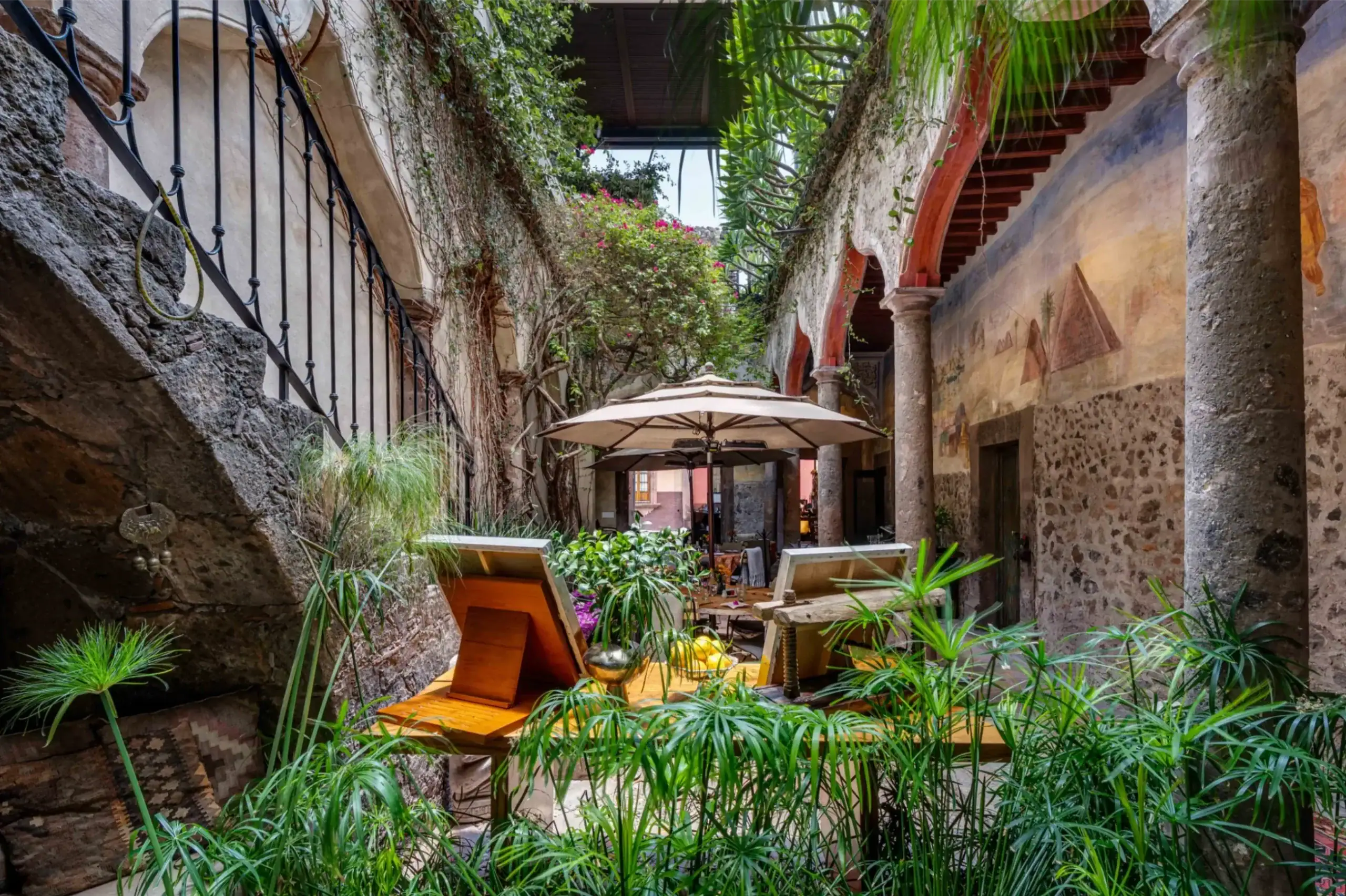 Stone-walled courtyard with arches, lush plants, and a wooden dining table shaded by a beige umbrella under a pergola collectives, San Miguel de Allende