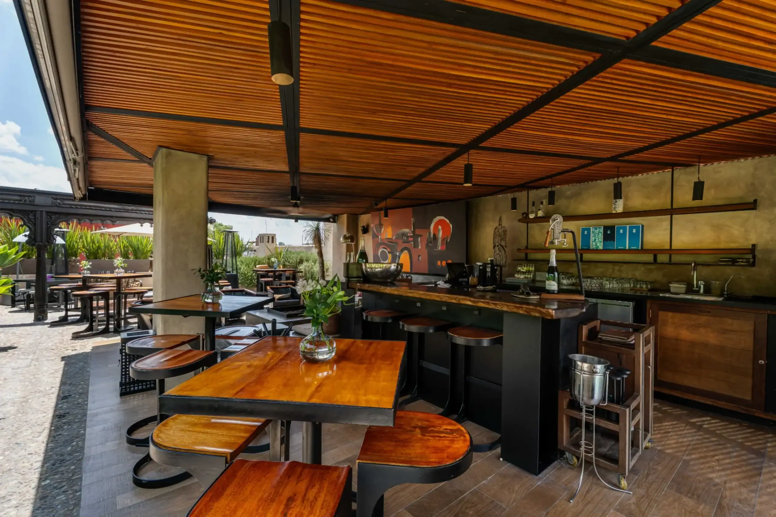 Open-air bar or cafe with long wooden tables and stools under a slatted wooden ceiling; plants and a mural are visible in the space, San Miguel de Allende