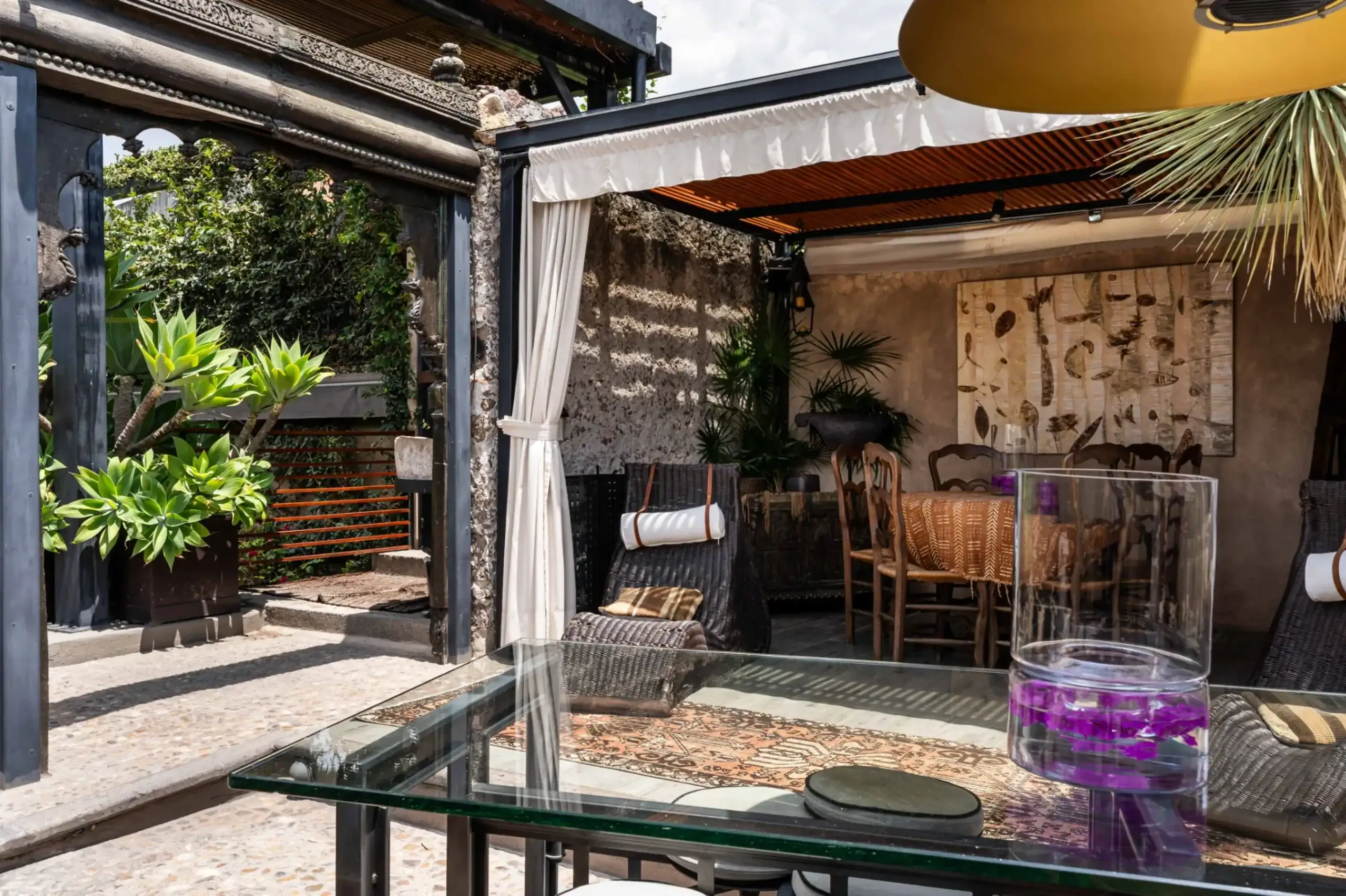 Outdoor seating area with a glass-top table, wicker chairs, and lush green plants under a shaded pergola, San Miguel de Allende