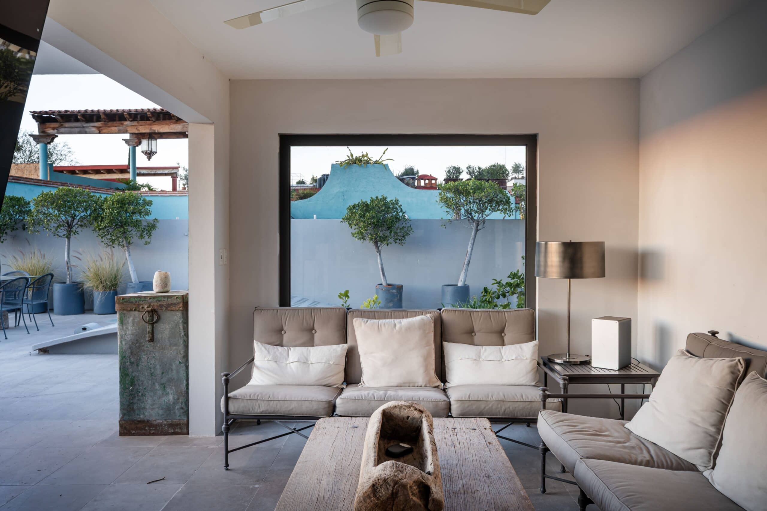 Indoor living room with beige sectional sofa facing a large window to a patio with blue wall and trees outside. A rustic wooden coffee table sits in front with a carved log centerpiece, San Miguel de Allende