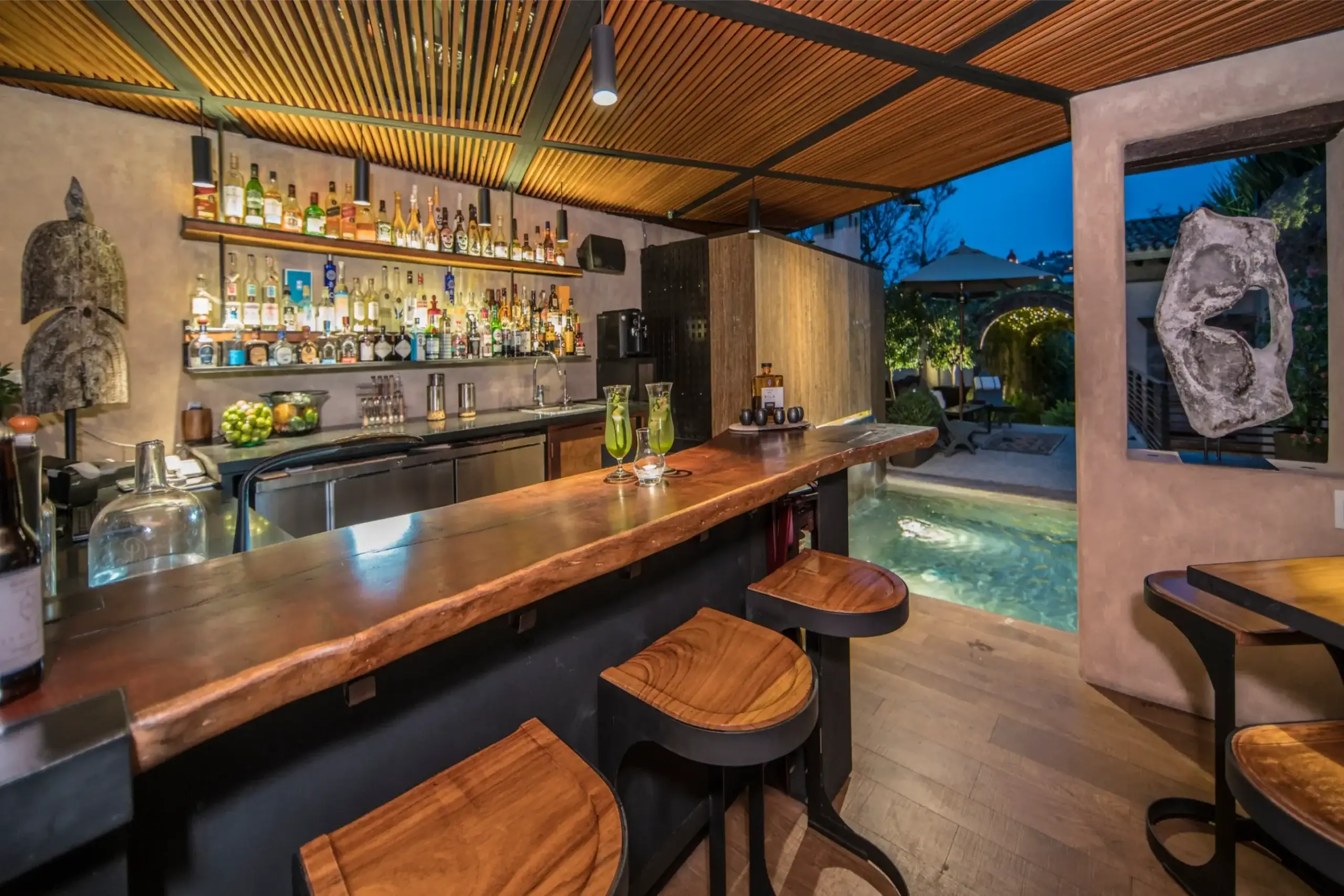 Bar area with a long wooden counter and matching stools, liquor bottles on backlit shelves, and warm pendant lights above, San Miguel de Allende