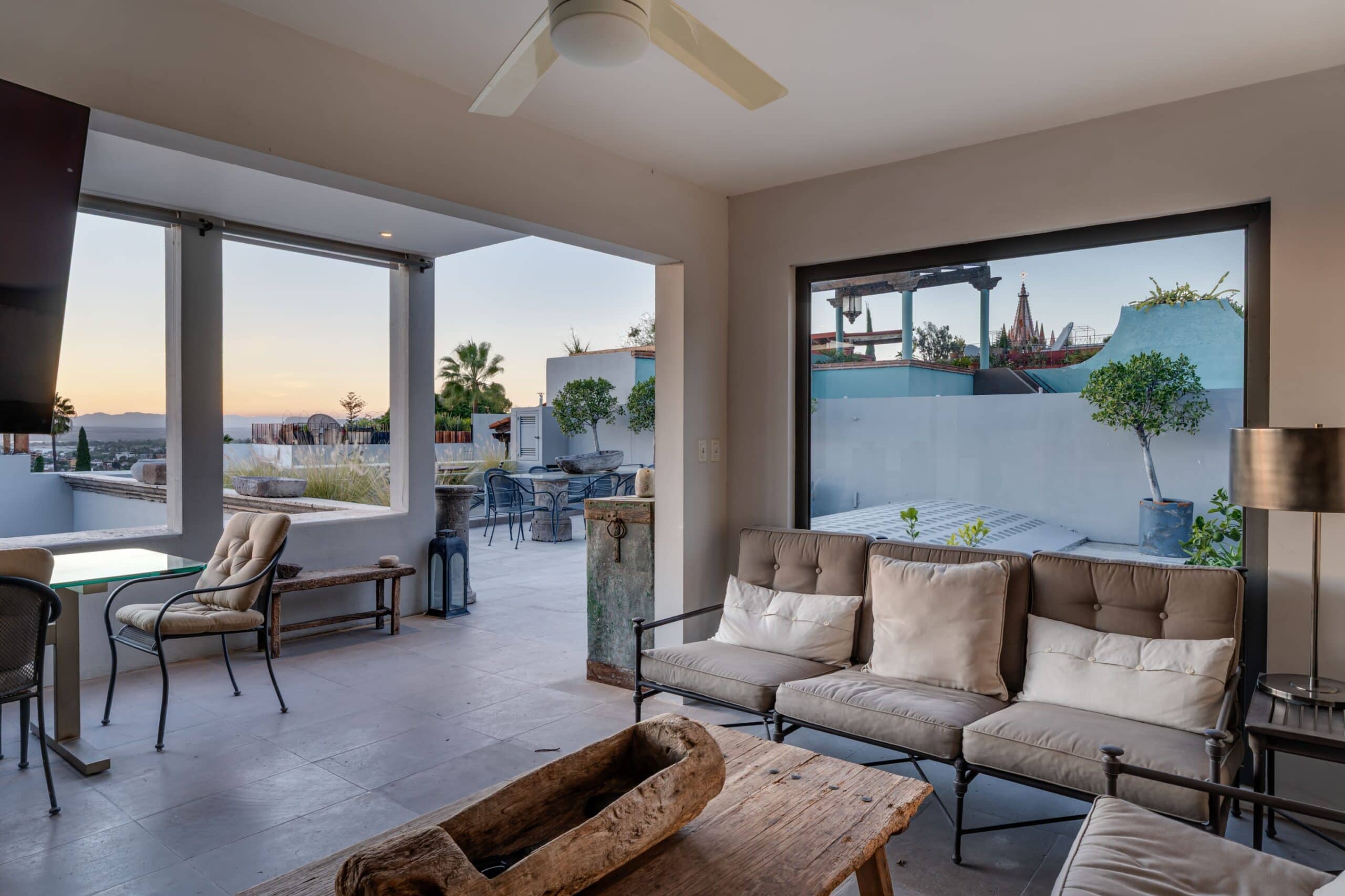 Lounge area with beige cushioned seating and a wooden coffee table, open to a sunny outdoor terrace viewed through large windows at sunset, San Miguel de Allende