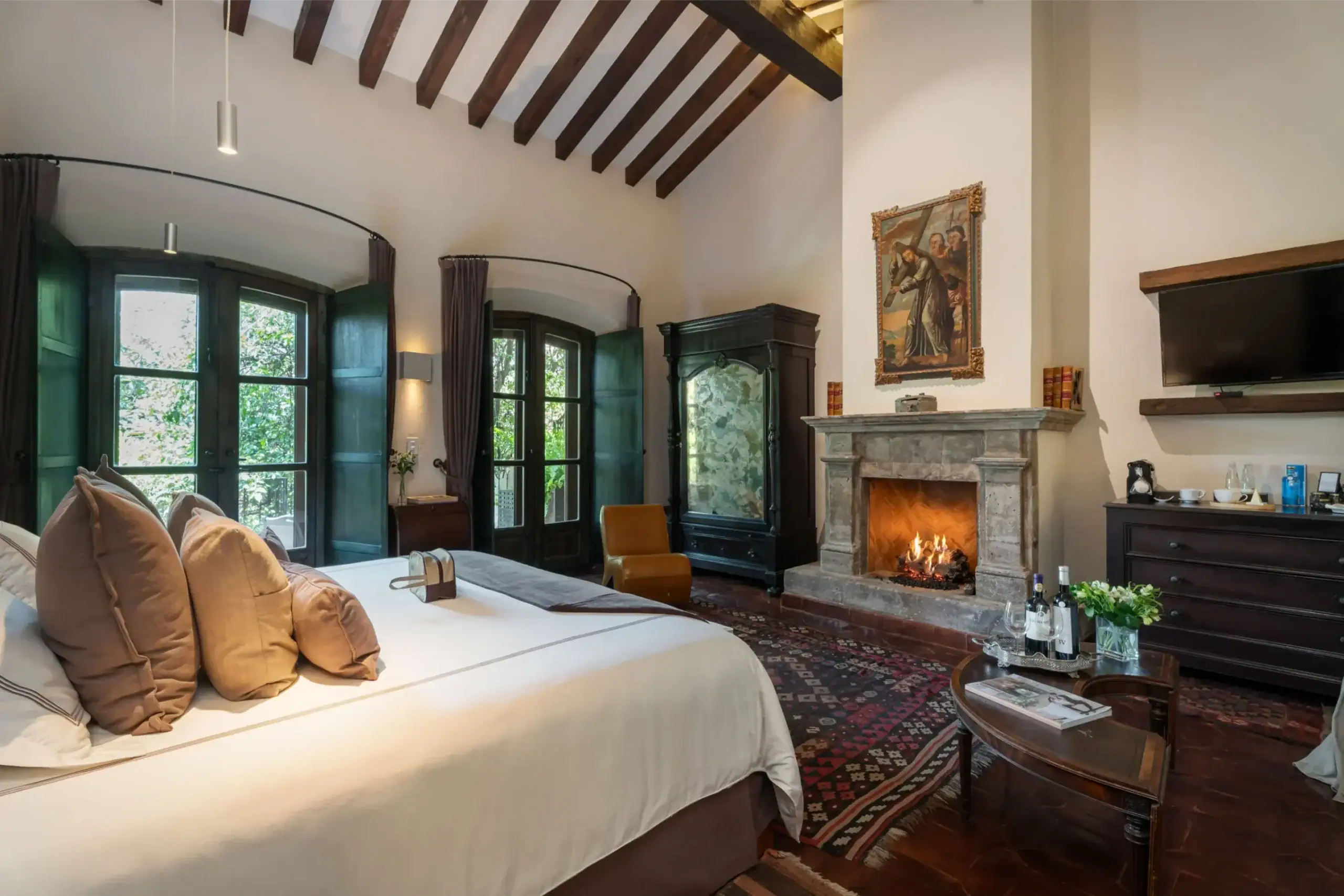 Luxurious bedroom with a lit stone fireplace, exposed wooden beams, and a large bed with tan cushions, San Miguel de Allende