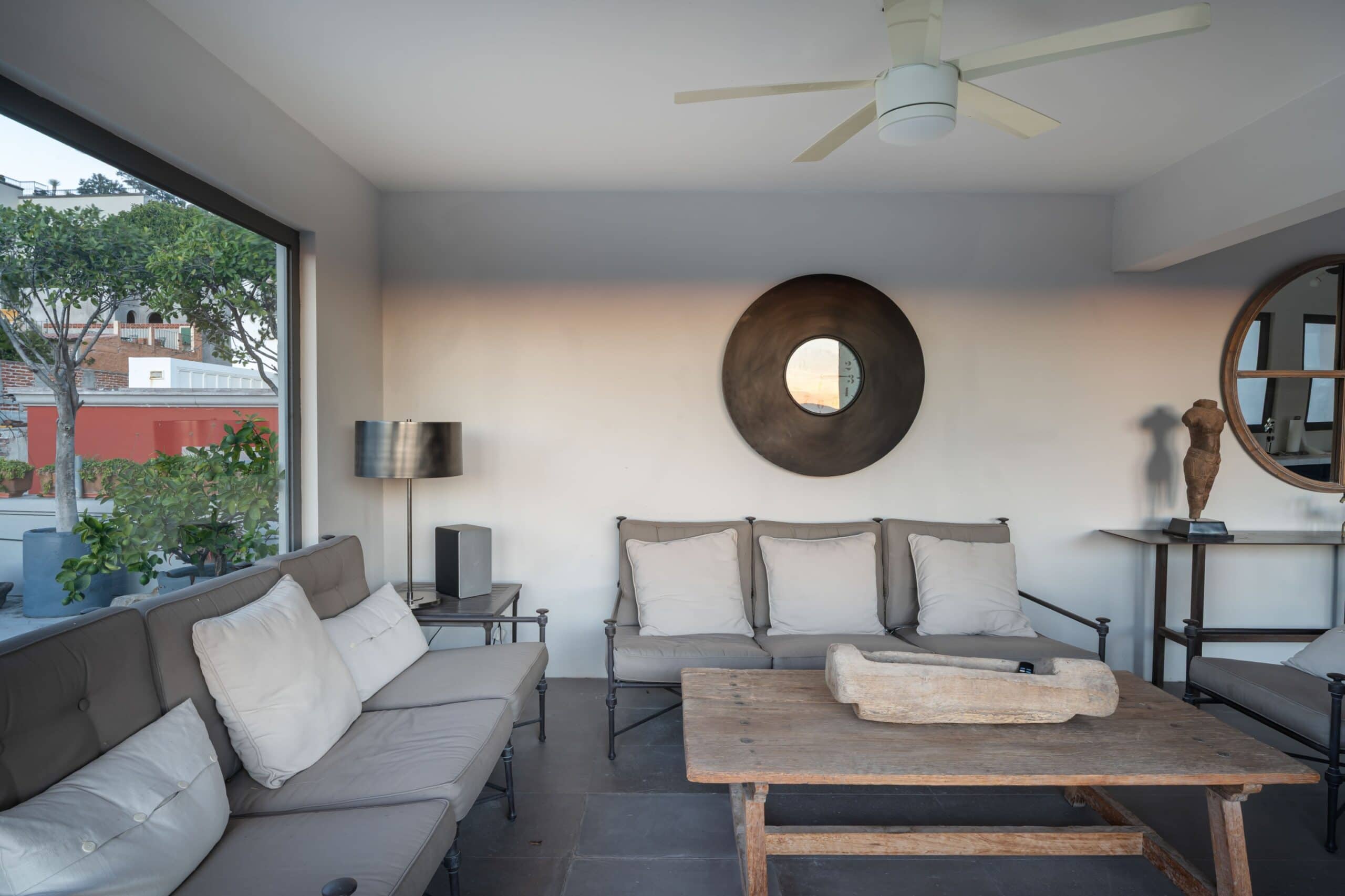 Modern living room interior with gray sectional sofa and white cushions, large window showing greenery outside, and a round black wall mirror, San Miguel de Allende