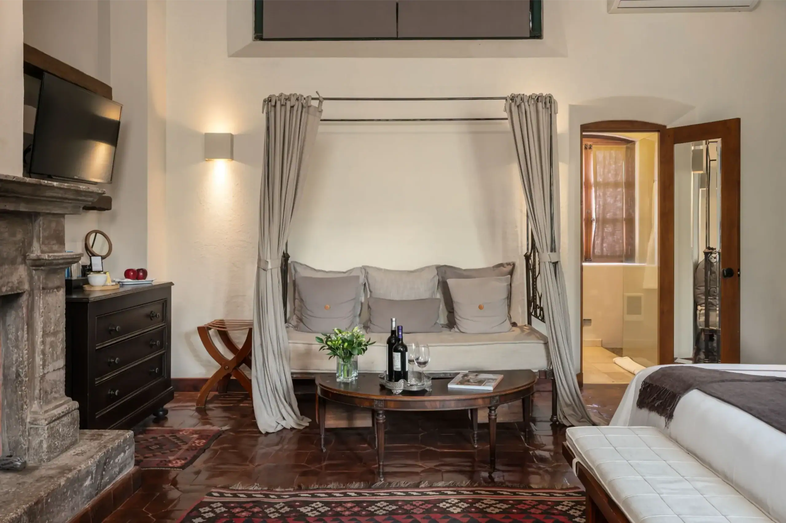 Cozy hotel sitting area with a cushioned four-poster daybed, gray curtains, and a round coffee table set with wine and glasses by the bed area, San Miguel de Allende