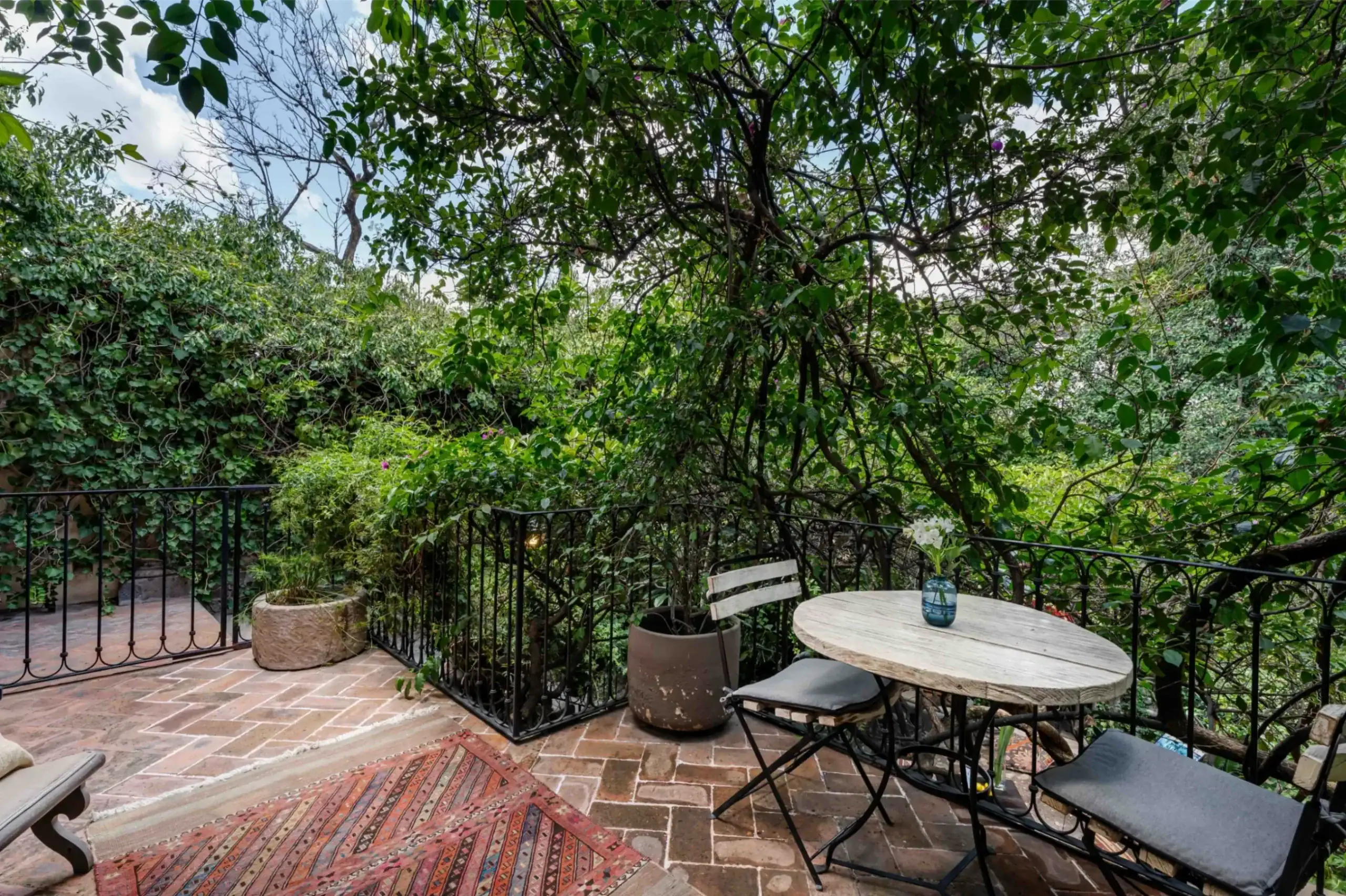 Outdoor brick patio with a round table and black metal chairs, surrounded by dense green vines and trees, San Miguel de Allende