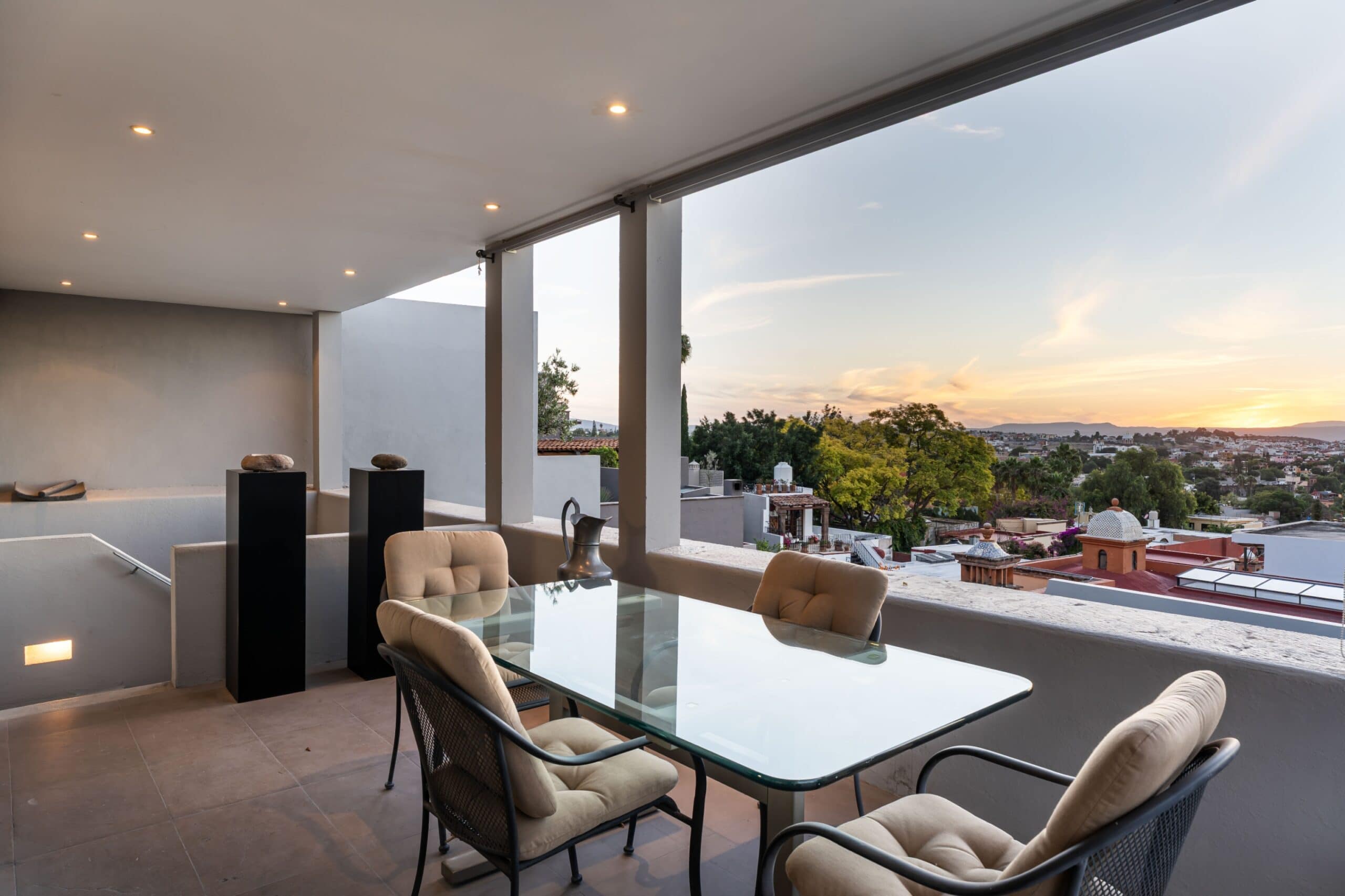 Covered balcony with a glass outdoor dining table and beige cushioned chairs, overlooking a city at sunset, San Miguel de Allende