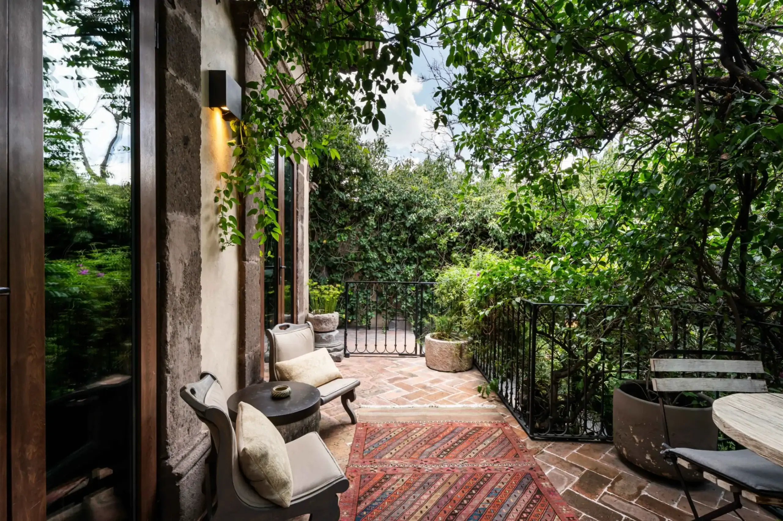 Outdoor patio with terracotta brick flooring, cushioned chairs, a round table, and a red patterned rug beside a stone wall covered in vines under a leafy canopy, San Miguel de Allende
