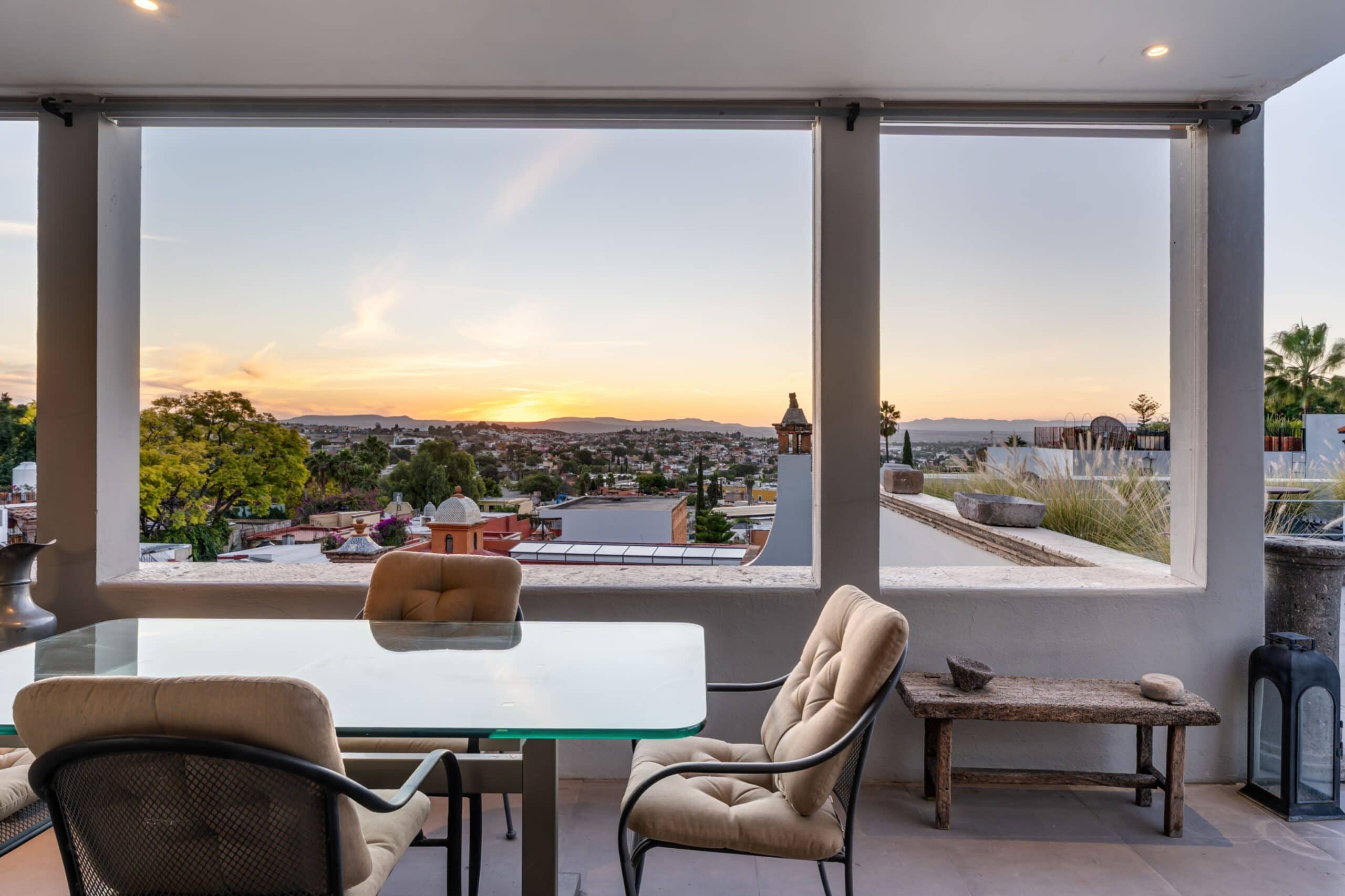 Outdoor balcony seating with a glass table and cushioned chairs, overlooking a sunlit town at sunset framed by hills in the distance, San Miguel de Allende