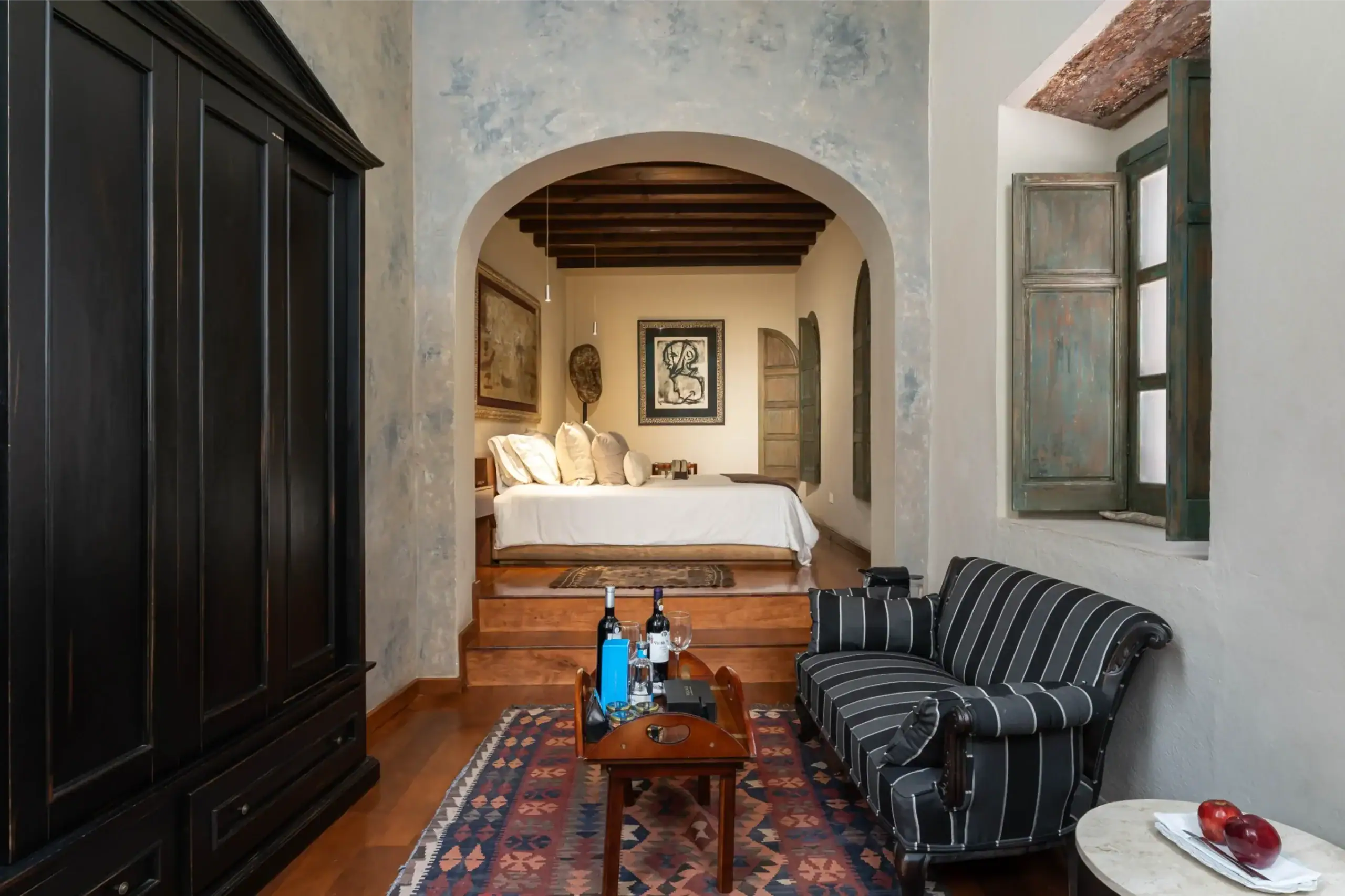 Dark wooden wardrobe on the left; an arched opening leads to a bedroom with a white bed and framed art, San Miguel de Allende