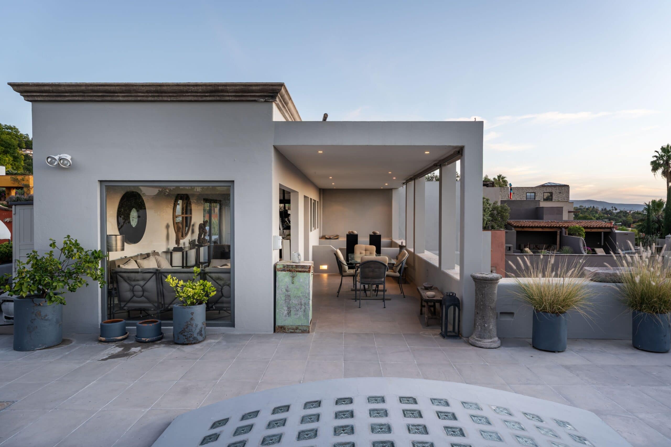 Modern outdoor patio on a gray stucco house with seating, potted plants, and an open covered dining area against a clear blue sky, San Miguel de Allende