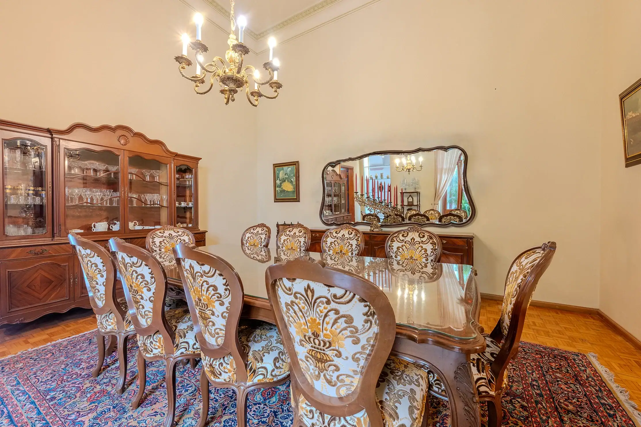 Elegant dining room with carved wooden chairs around a glass table, a china cabinet, chandelier, and mirror in the background, San Miguel de Allende