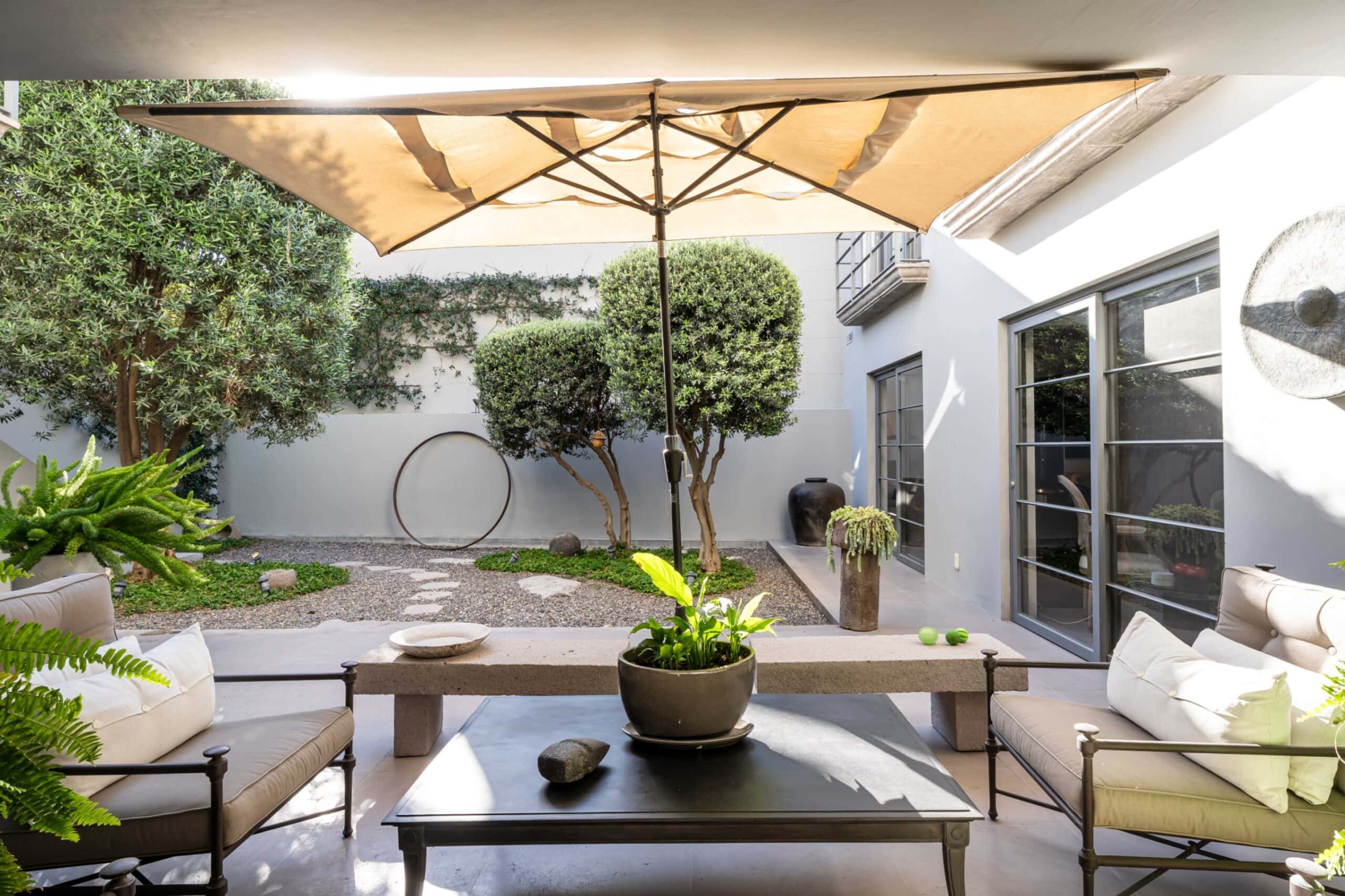 Outdoor patio with beige cushioned seating around a dark coffee table under a large beige umbrella, surrounded by greenery and glass doors, San Miguel de Allende