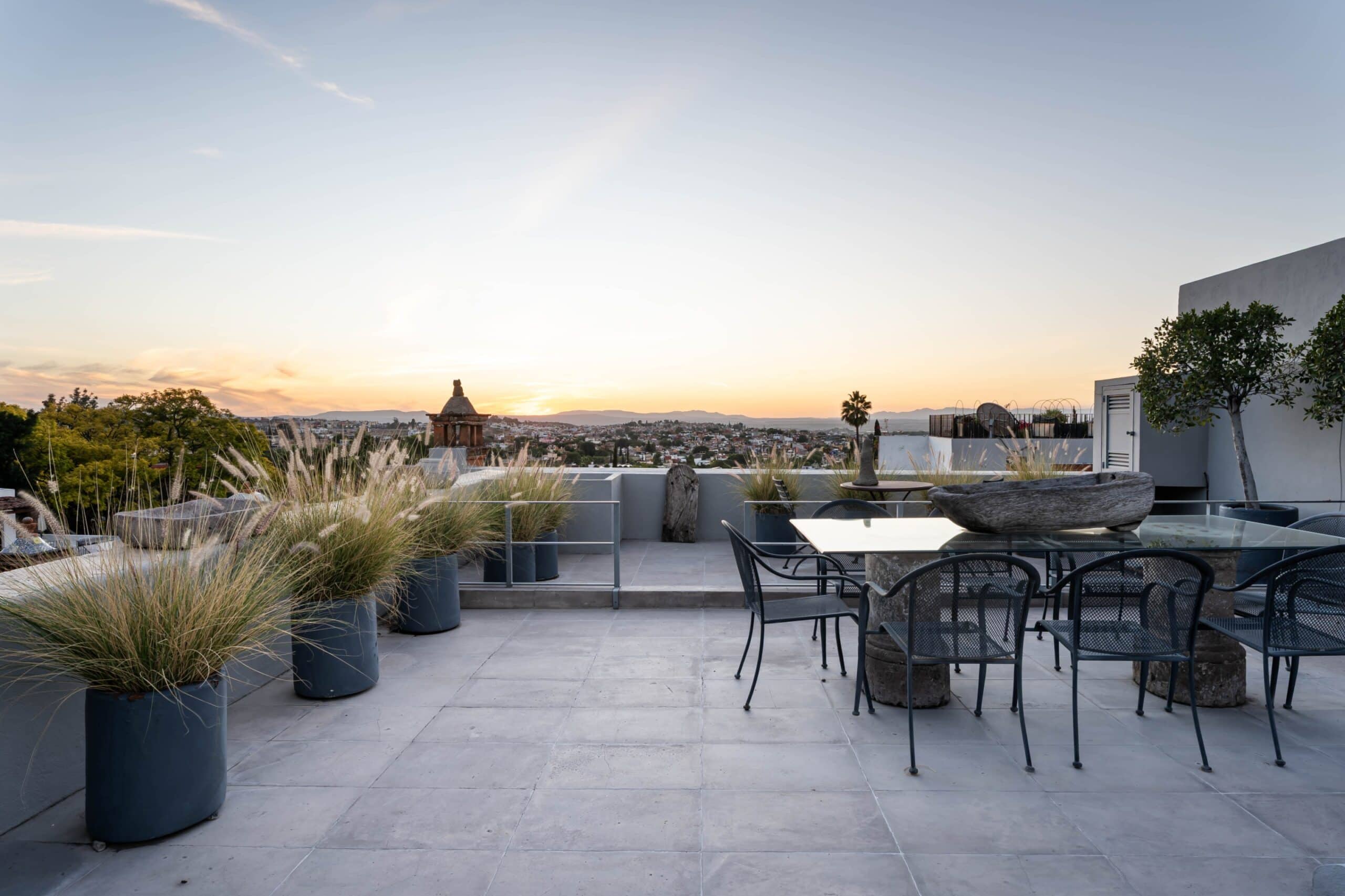 Rooftop terrace at sunset with outdoor dining tables and chairs, potted grasses, and a distant cityscape, San Miguel de Allende