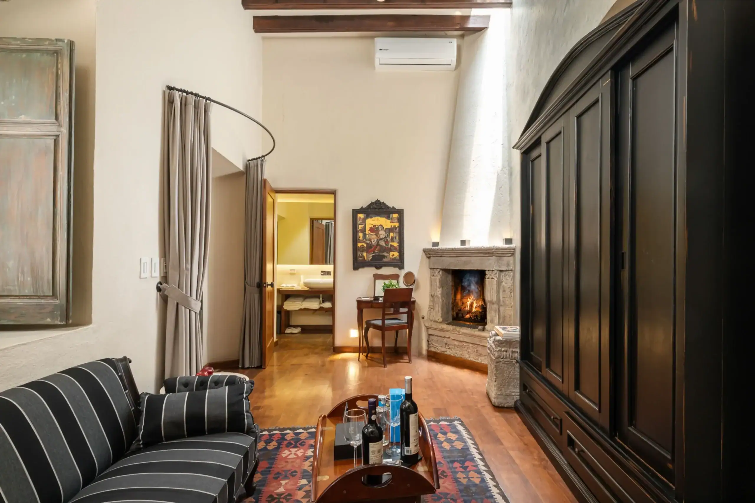 Cozy living room with a stone fireplace glowing, dark wood built-in cabinets, and a striped black-and-gray sofa facing a wooden coffee table with wine bottles, San Miguel de Allende
