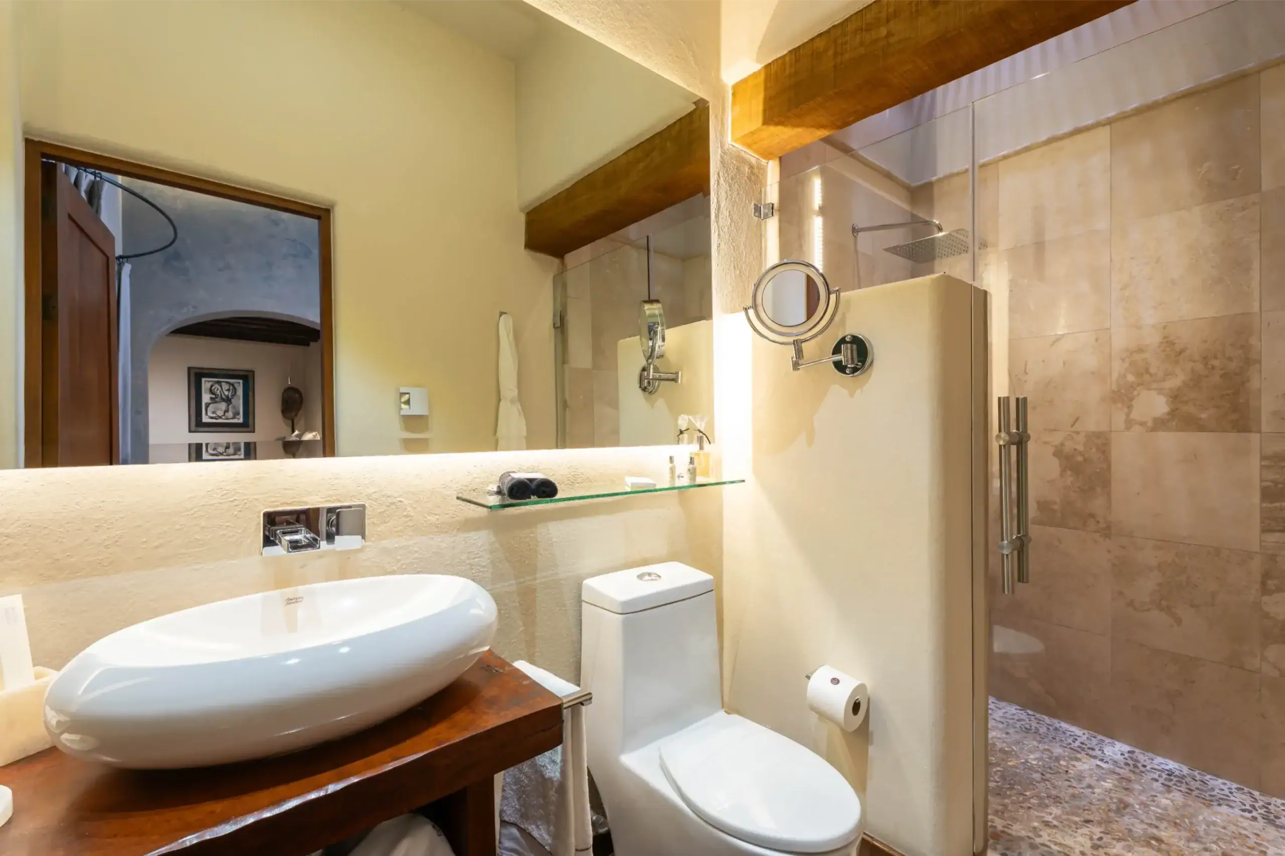 Modern bathroom with a white oval vessel sink on a wooden vanity, mirrored wall, and glass shower enclosure, San Miguel de Allende