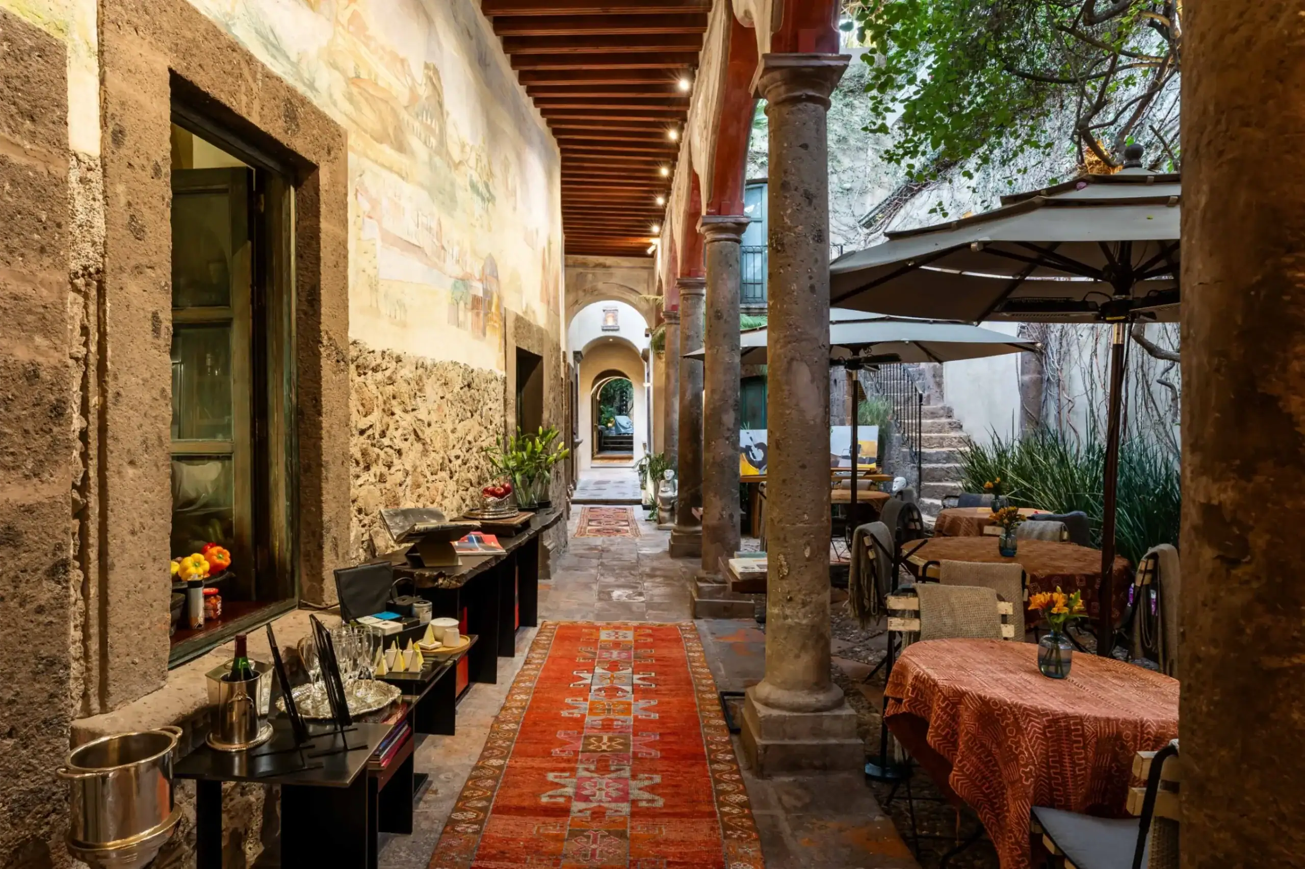 Narrow stone courtyard cafe with tall columns, arches, and a red patterned rug along tables set with flowers under string lights, San Miguel de Allende