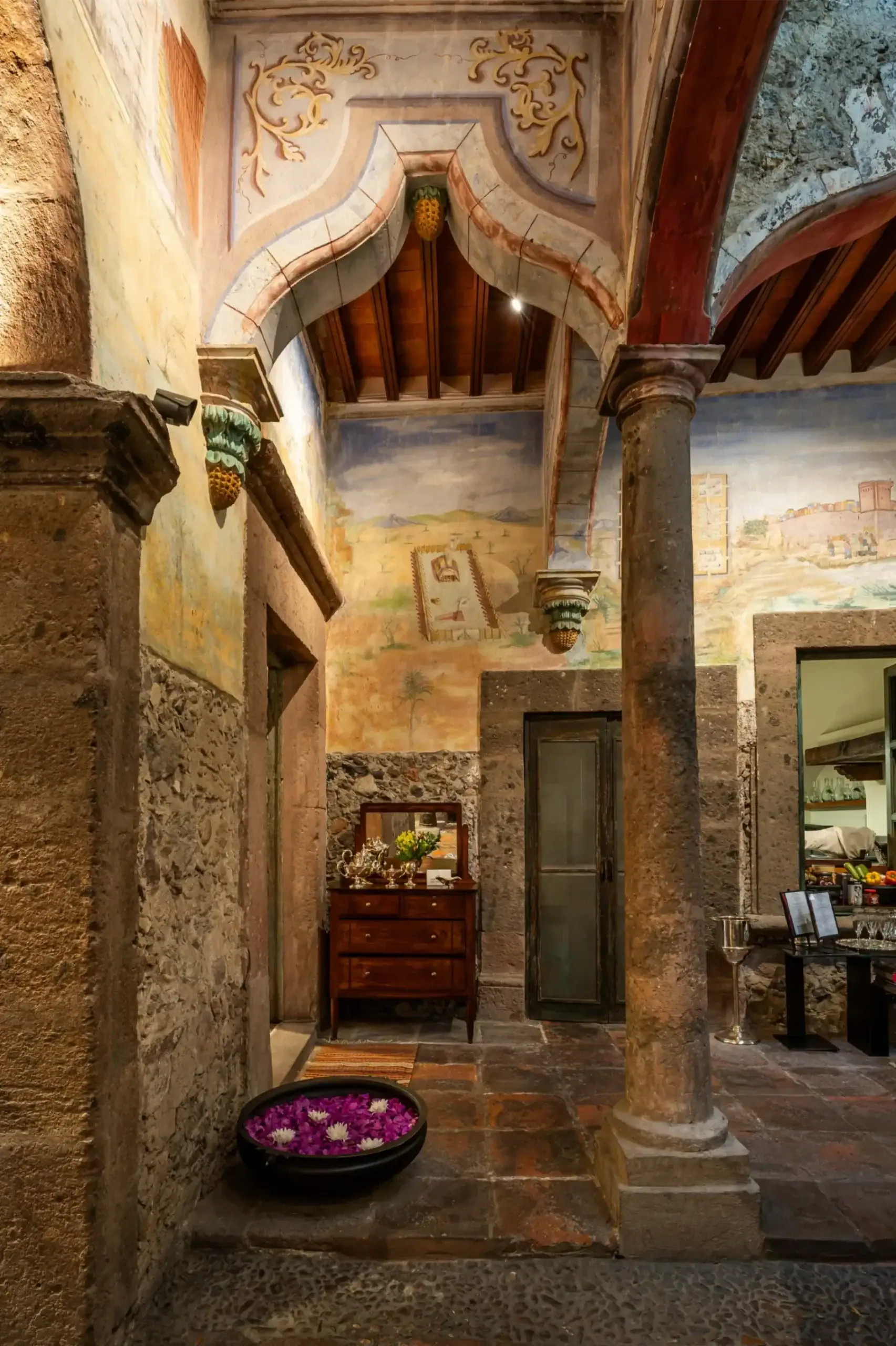 Interior of a historic courtyard with ornate arches and stone columns, painted walls, and a wooden dresser displaying silverware nearby, San Miguel de Allende