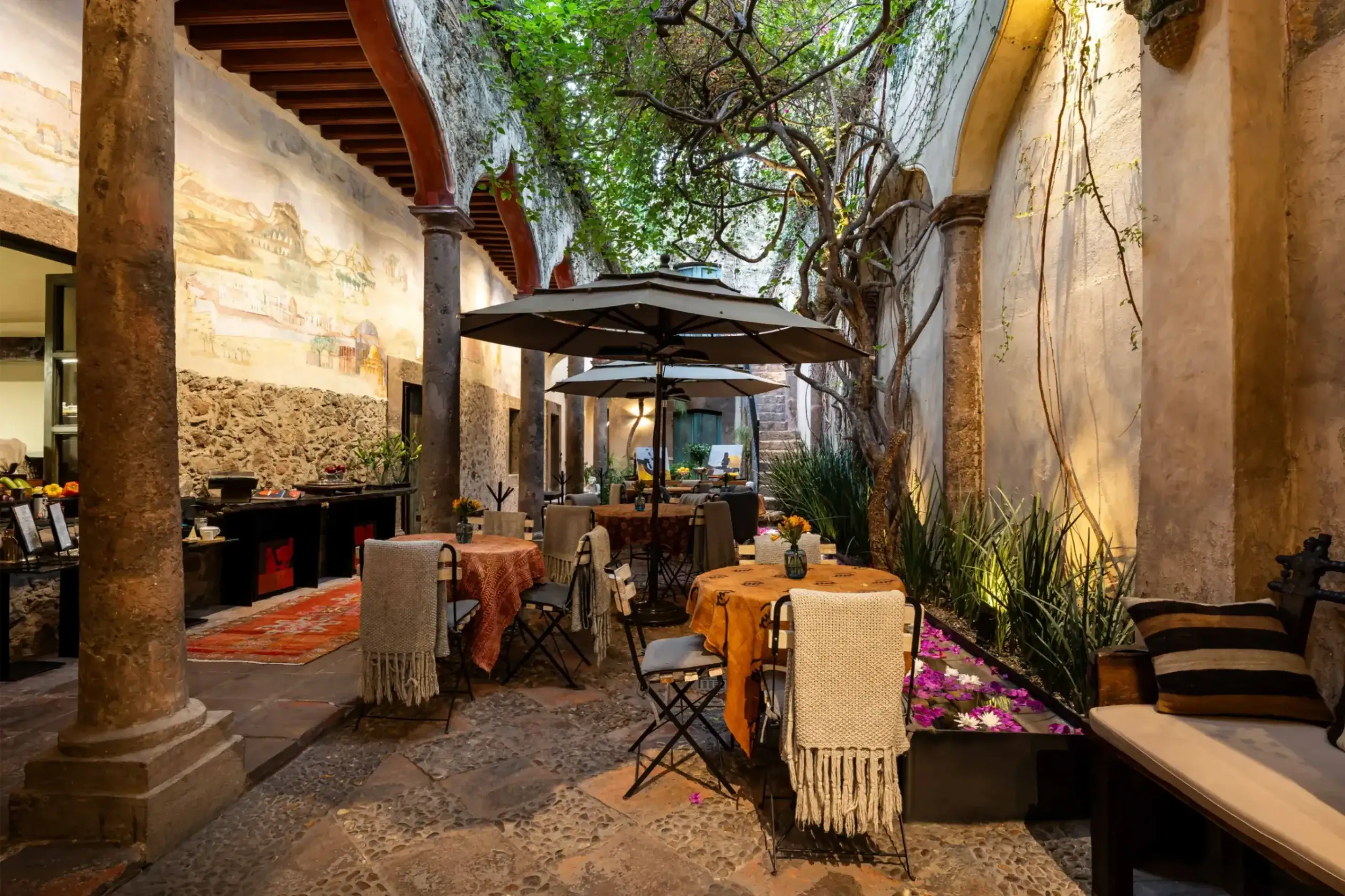 Outdoor courtyard dining area with tables covered in orange tablecloths, beige woven chairs, and large umbrellas under stone columns and vines on the walls, San Miguel de Allende