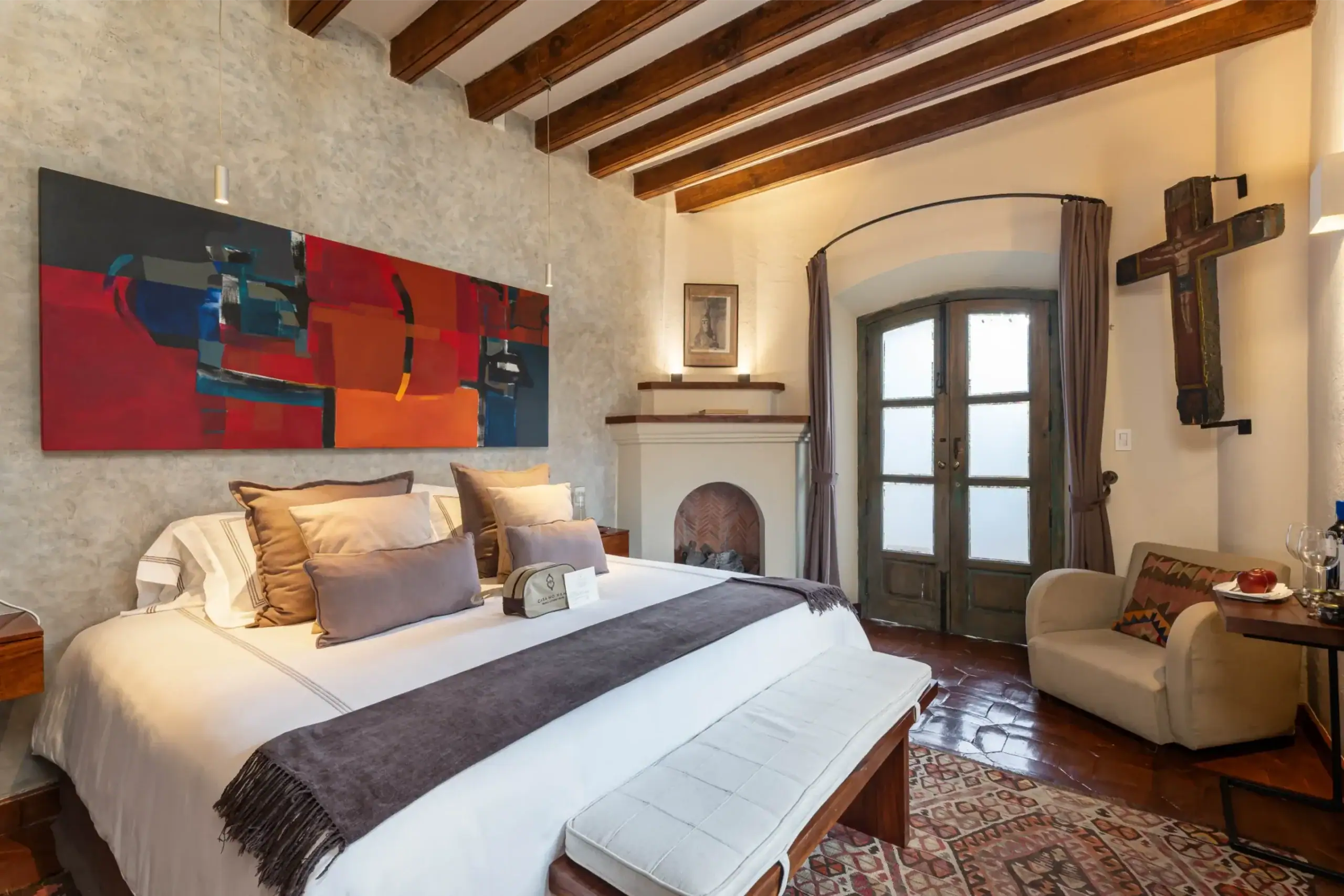 Cozy hotel room with a large bed, colorful abstract painting, exposed wooden beams, and a stone fireplace wall, San Miguel de Allende