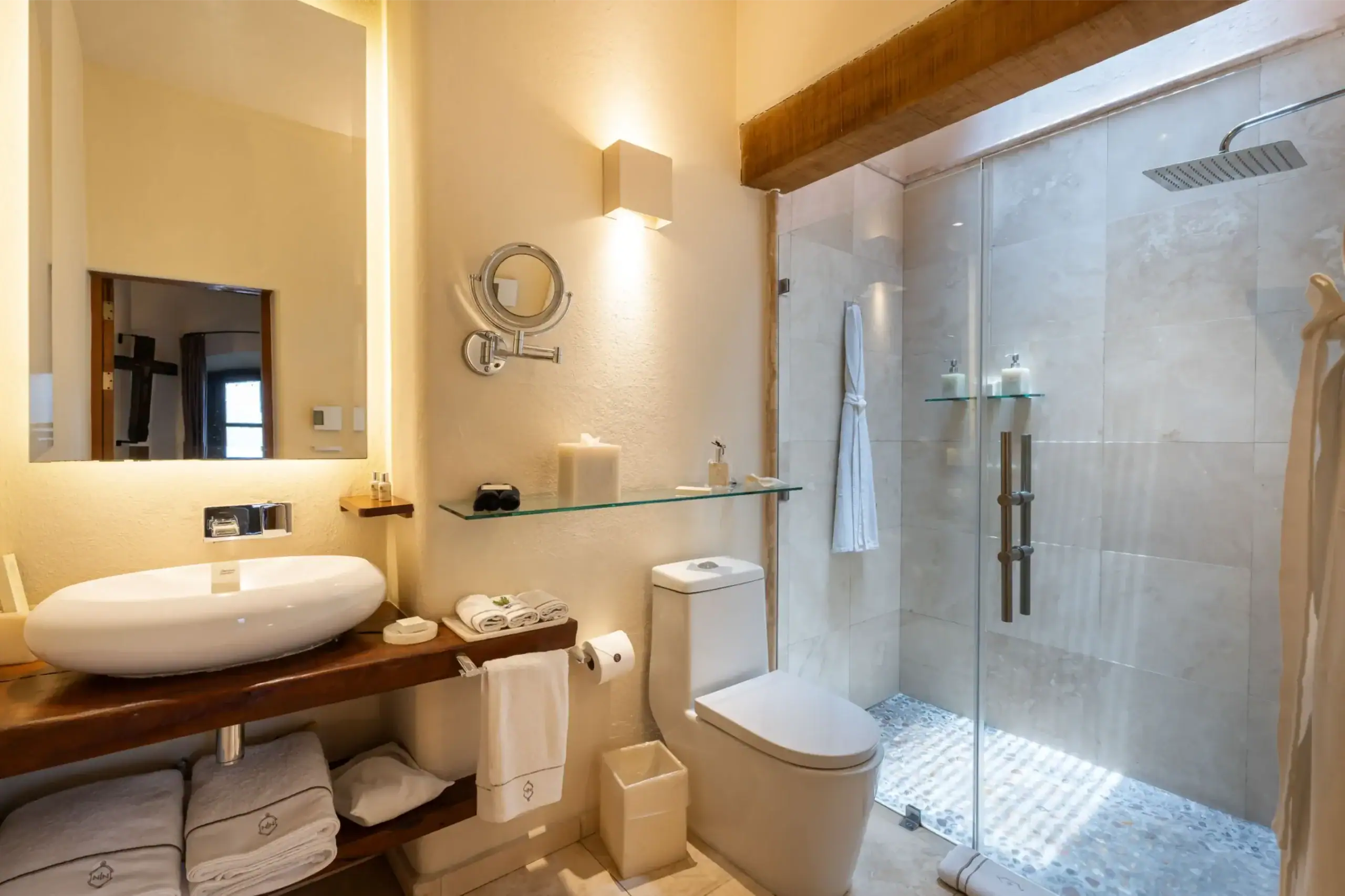 Modern bathroom with glass shower enclosure, rainfall showerhead, and white vessel sink on a wooden counter with folded towels nearby, San Miguel de Allende
