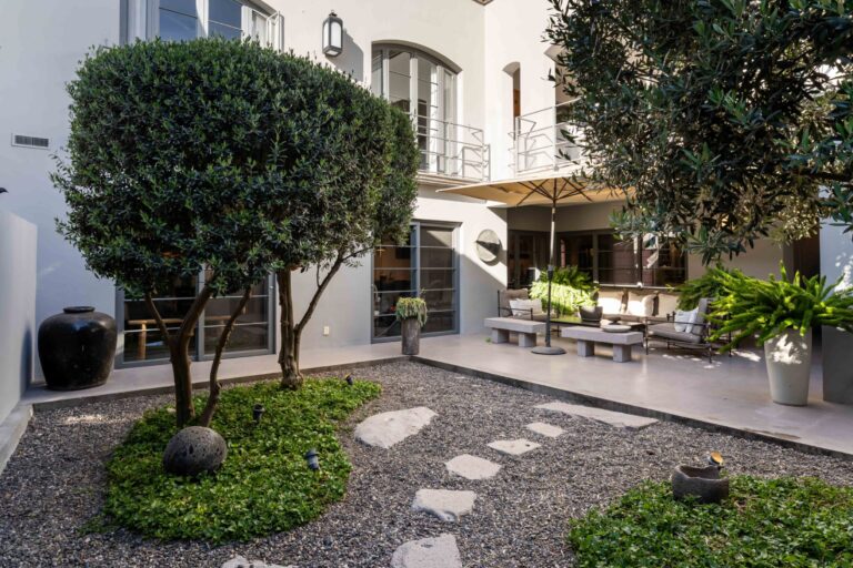 Modern courtyard with gravel yard, stepping stones, and two large trees; patio seating under a beige umbrella beside glass doors, San Miguel de Allende