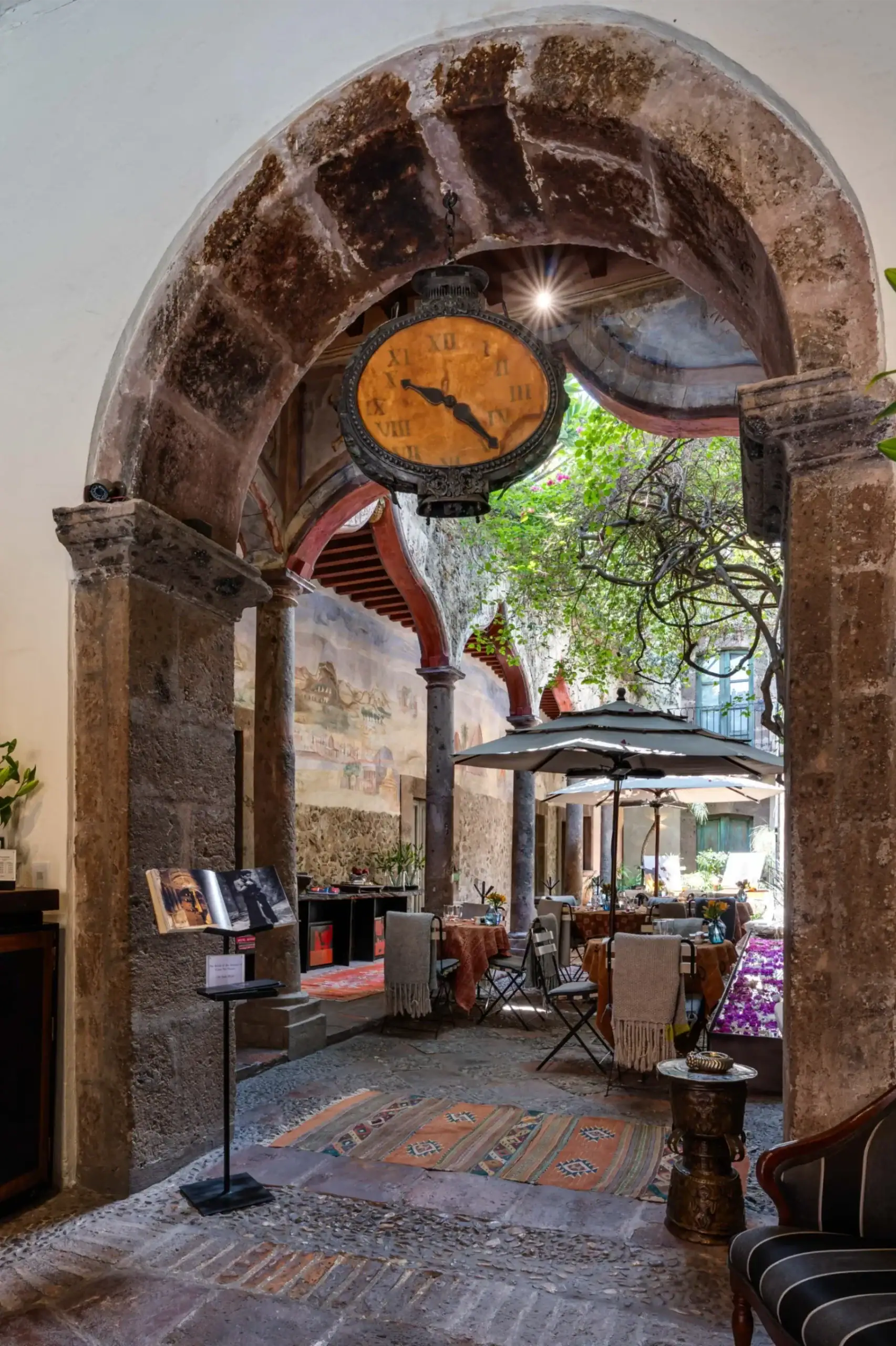 Stone archway cafe courtyard with an antique hanging clock, outdoor tables, and colorful rugs beneath, San Miguel de Allende