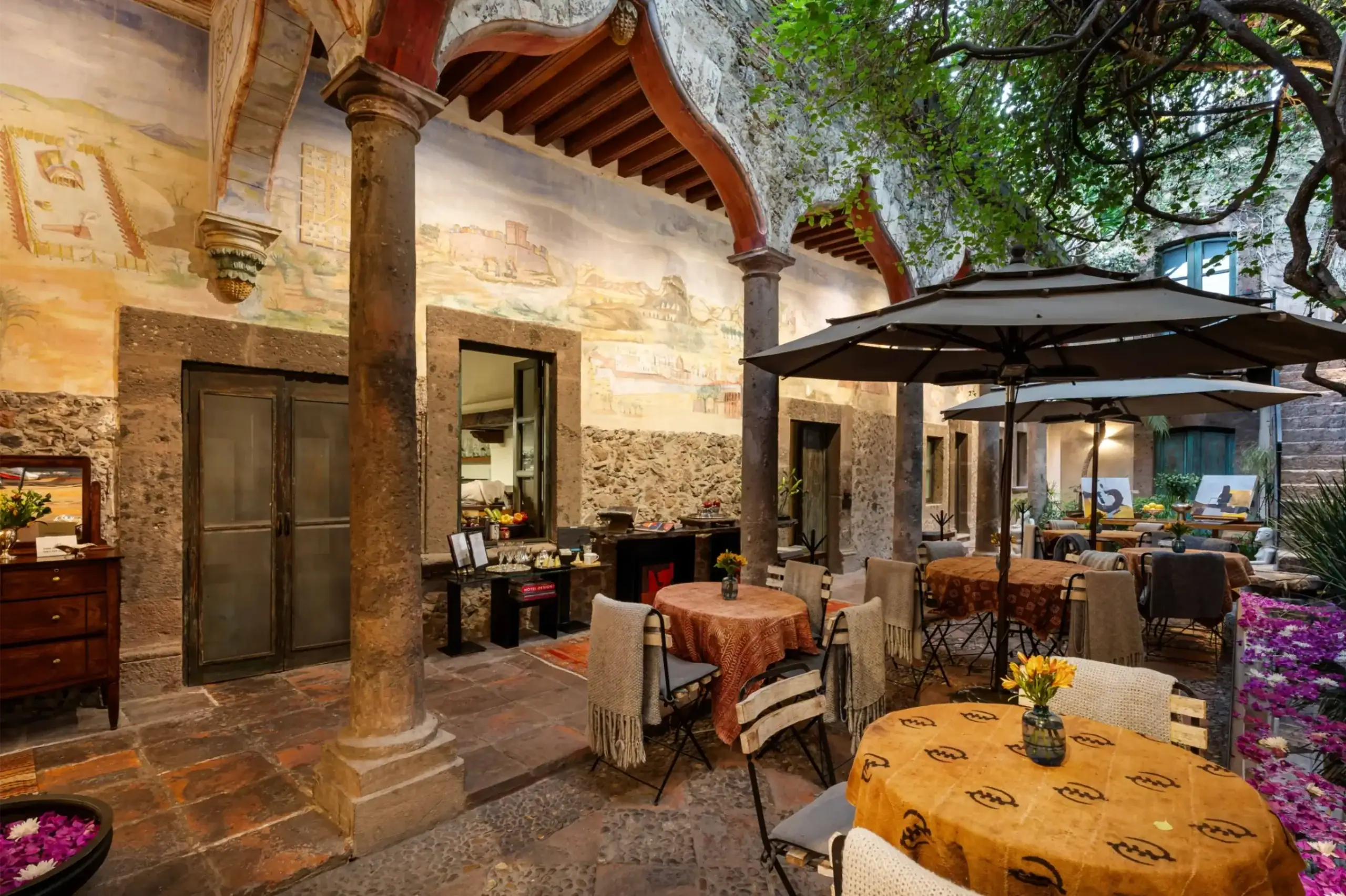 Outdoor dining courtyard with round tables covered in yellow cloths, wicker chairs, and large beige umbrellas beneath stone arches under a tree-filled canopy, San Miguel de Allende