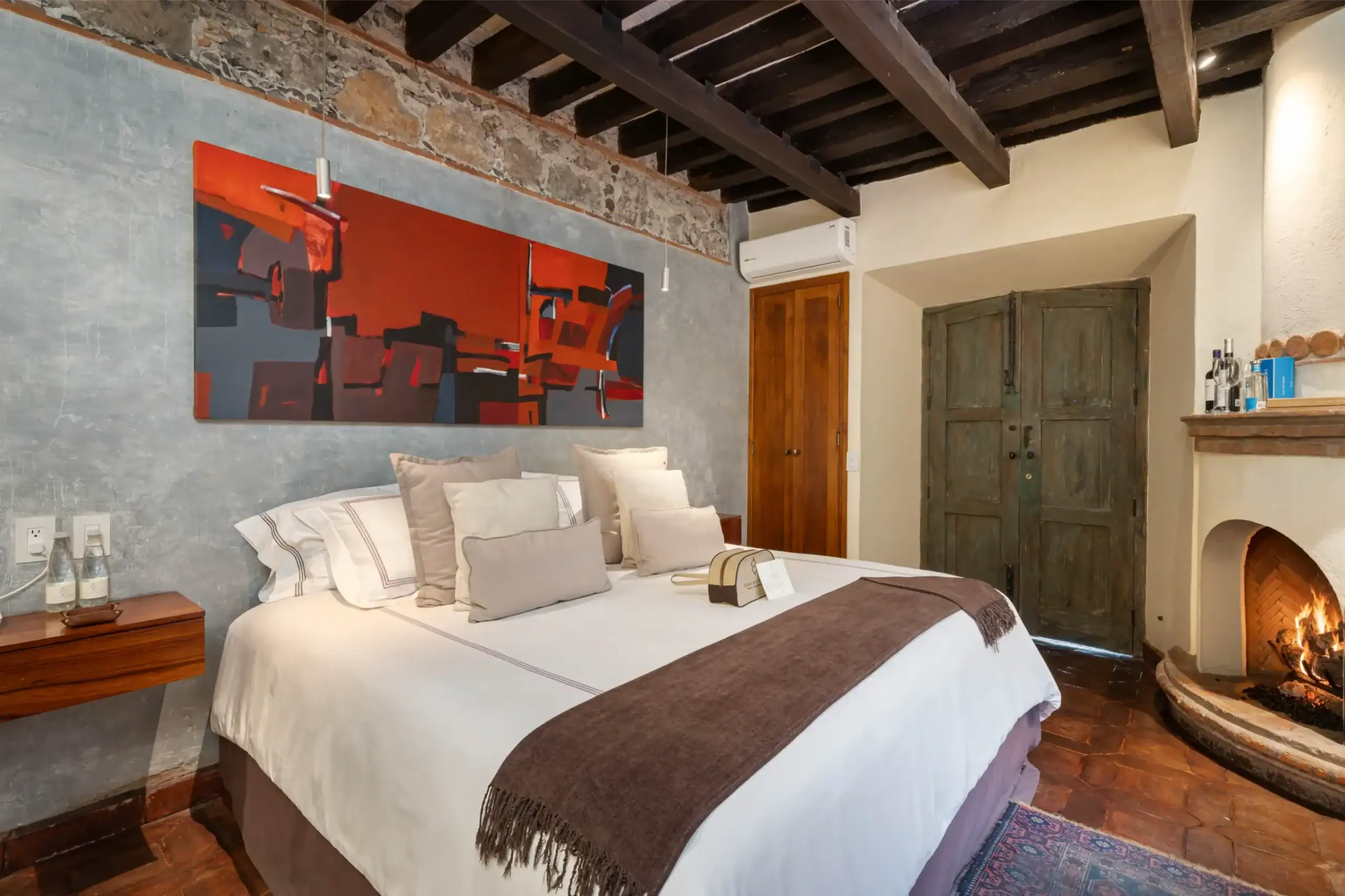 Rustic hotel bedroom with a large bed, neutral pillows, and a bold abstract painting on a gray textured wall under dark wooden beams, San Miguel de Allende