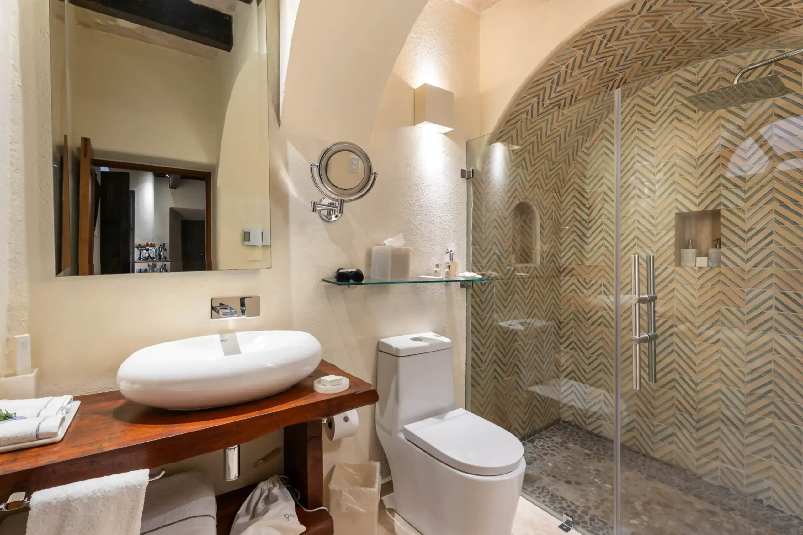 Modern bathroom with oval white vessel sink on a wooden vanity, mirrored wall, and round magnifying mirror above a glass shelf, San Miguel de Allende