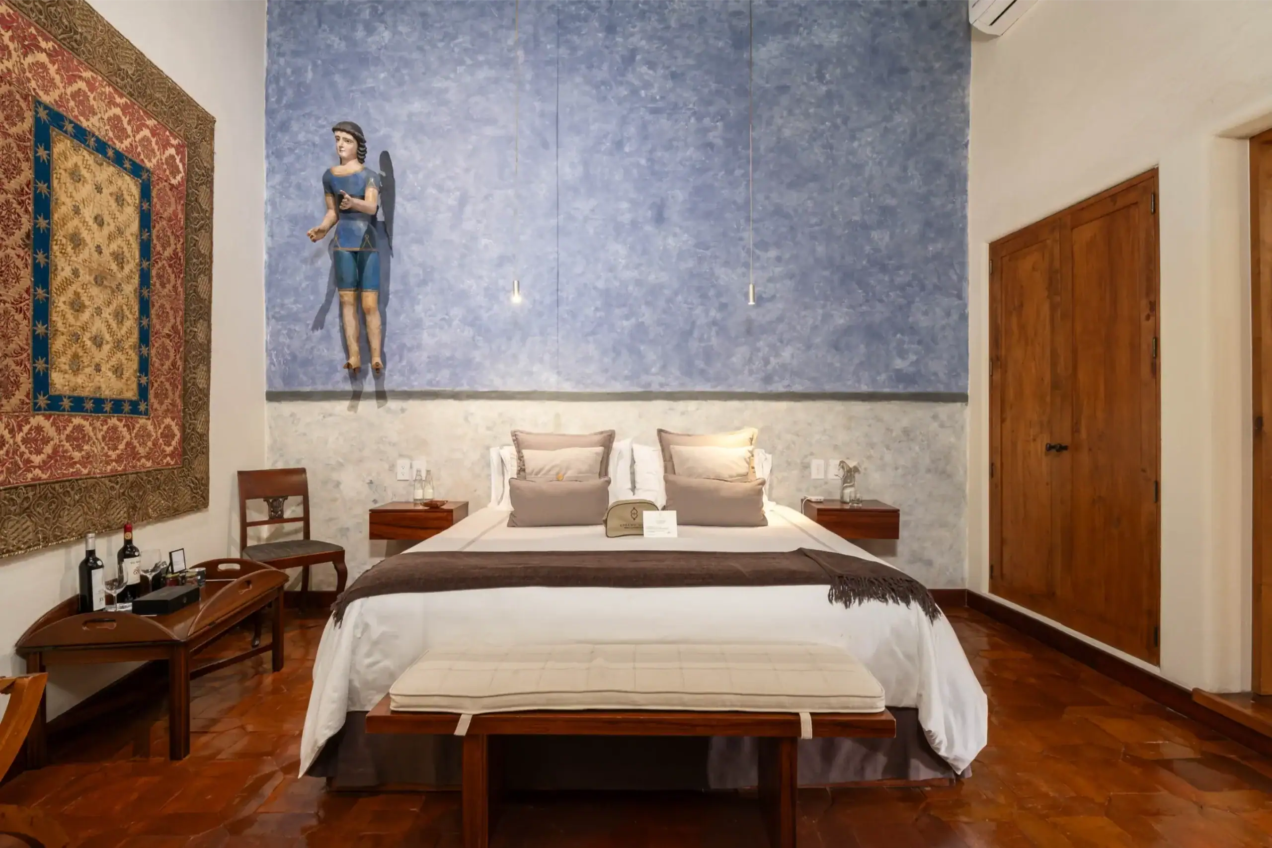 Hotel-style bedroom with a blue textured wall, hanging angel figure above the bed, and a wooden door to the right, San Miguel de Allende