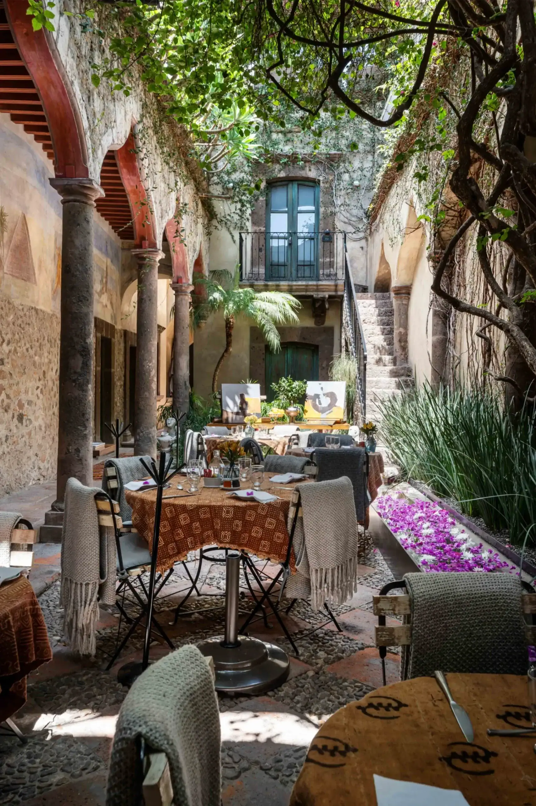 Outdoor courtyard restaurant with round tables, wicker chairs, and table settings under a vine-covered stone arcade, San Miguel de Allende