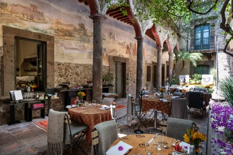 Outdoor courtyard restaurant with stone columns and red arches, long muraled wall, and tables set for dining under green foliage, San Miguel de Allende