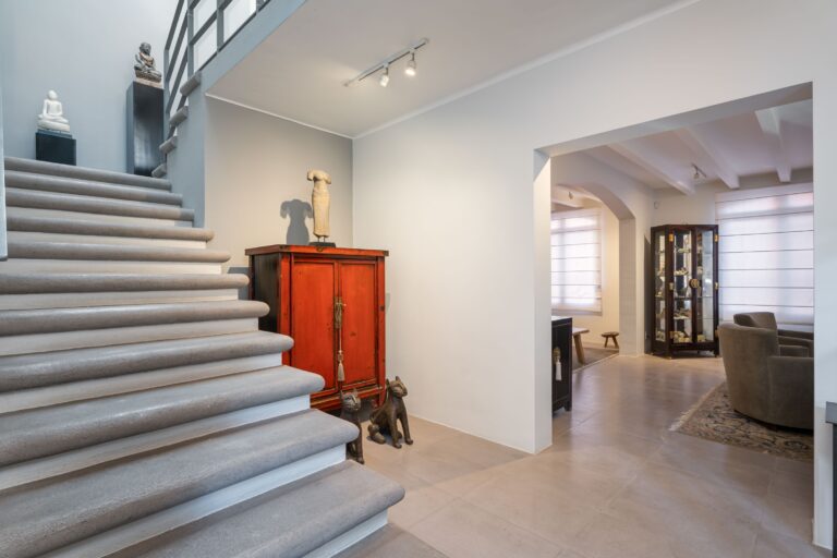 Interior of a modern living space: stone staircase on the left, statues on pedestals, a red antique cabinet, and a distant glass display cabinet through an archway, San Miguel de Allende