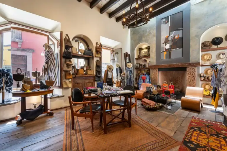 Boutique interior filled with handbags on shelves, hats on display, and wooden furniture under rustic beams and warm lighting, San Miguel de Allende
