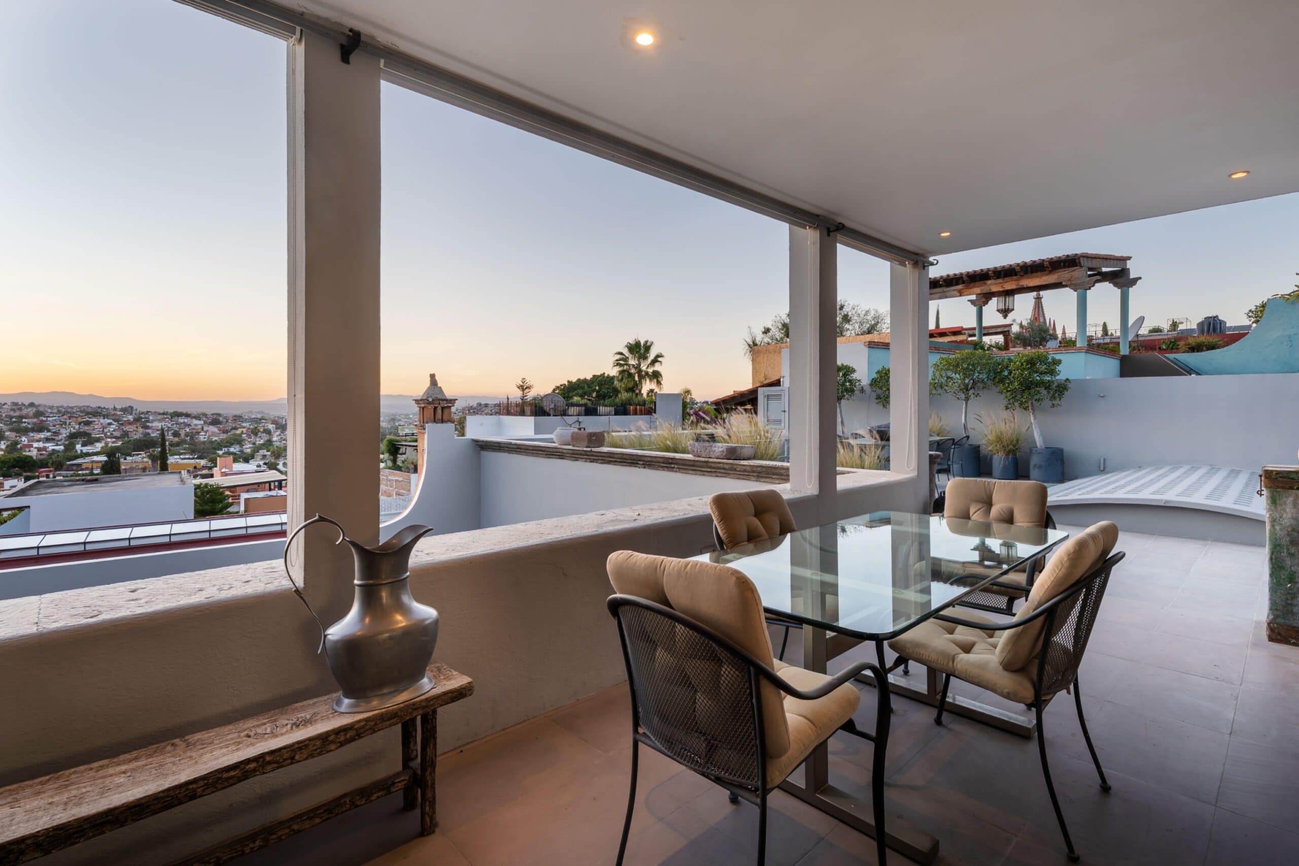 Rooftop patio with a glass dining table and tan cushioned chairs, overlooking a city at sunset, San Miguel de Allende