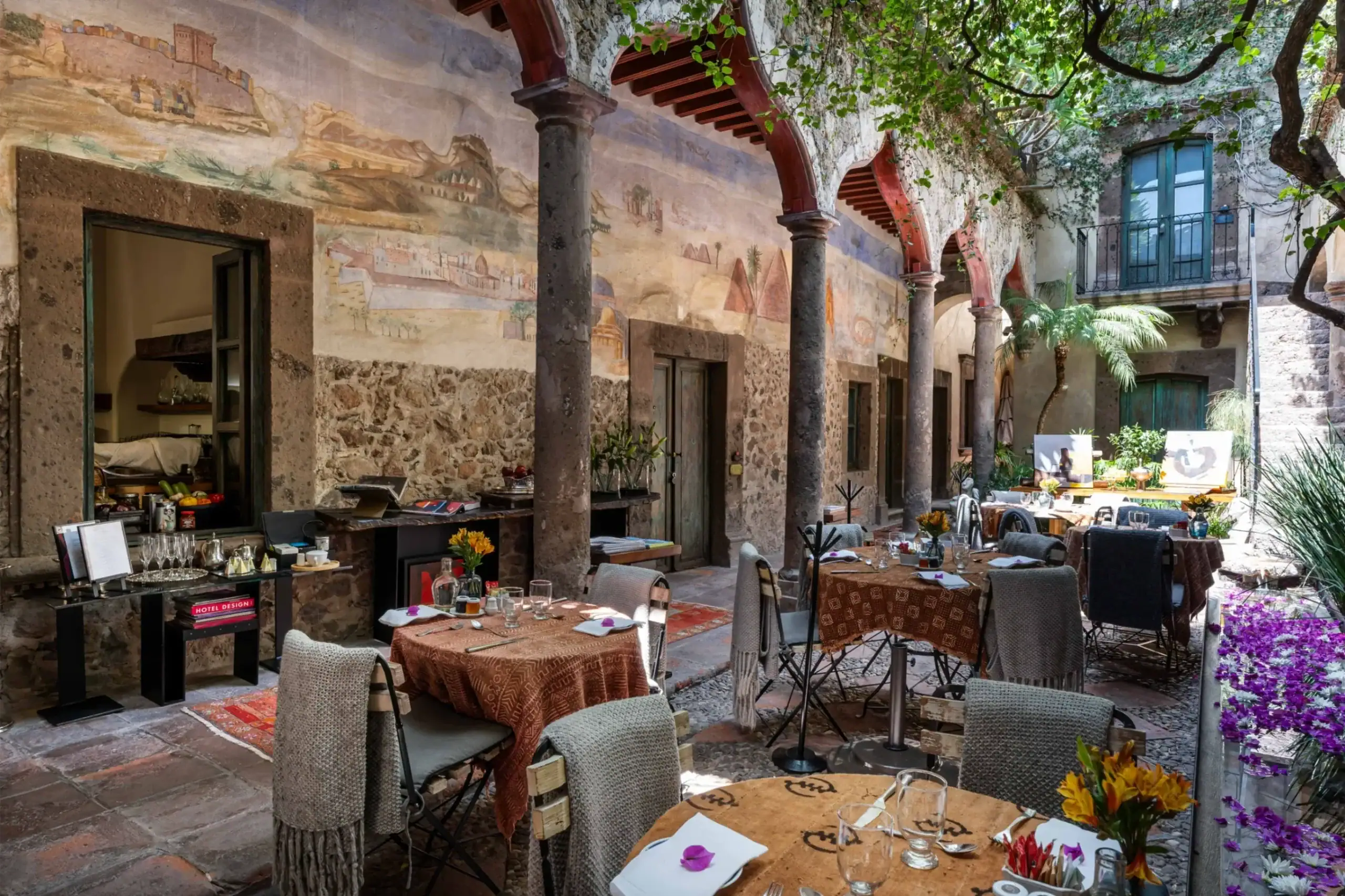 Outdoor courtyard restaurant with stone columns and red arches, long muraled wall, and tables set for dining under green foliage, San Miguel de Allende